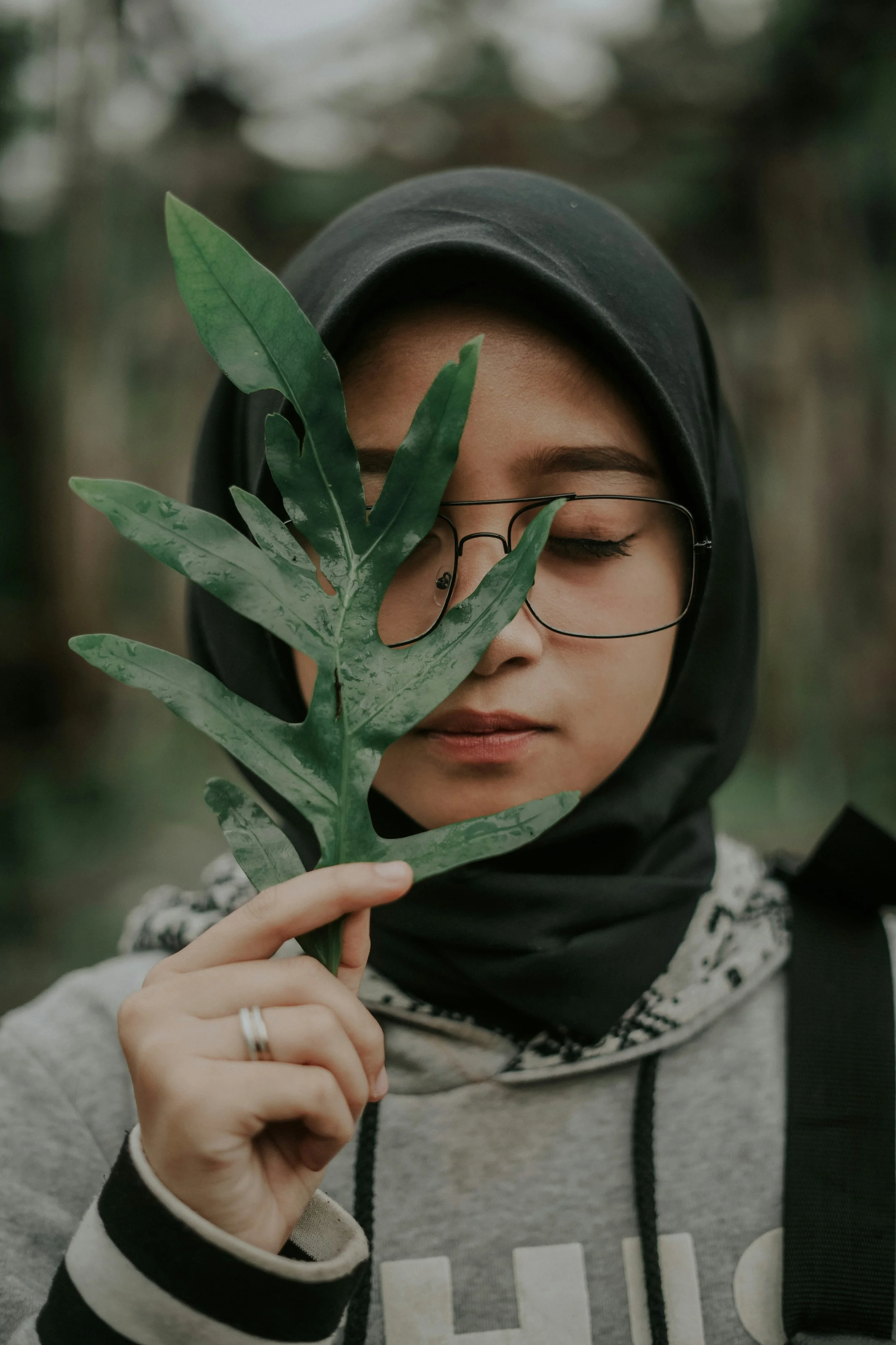 A woman wearing glasses and a hijab holding a green leaf in front of her face, with her eyes closed.