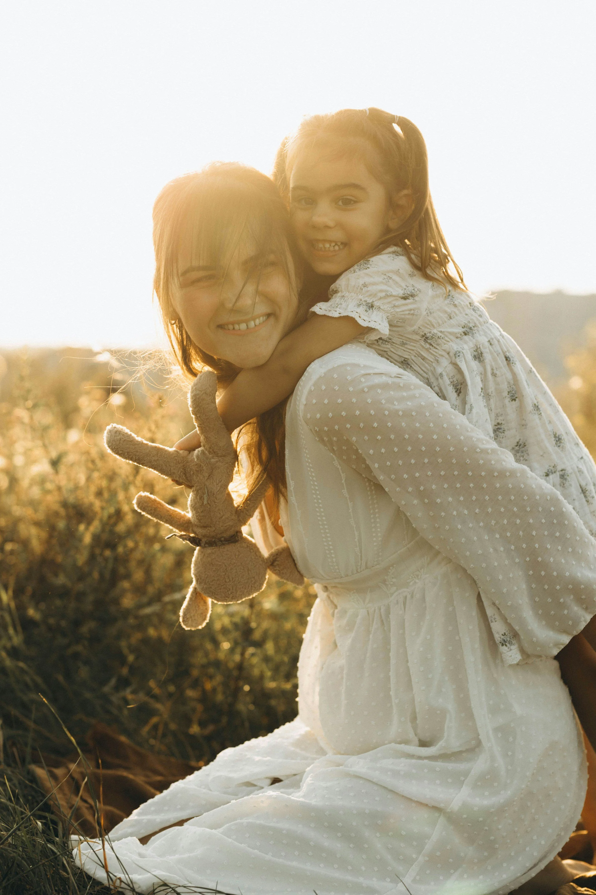A woman with red hair and glasses holds and embraces a young girl with pigtails and a white dress while sitting outdoors in a field at sunset.