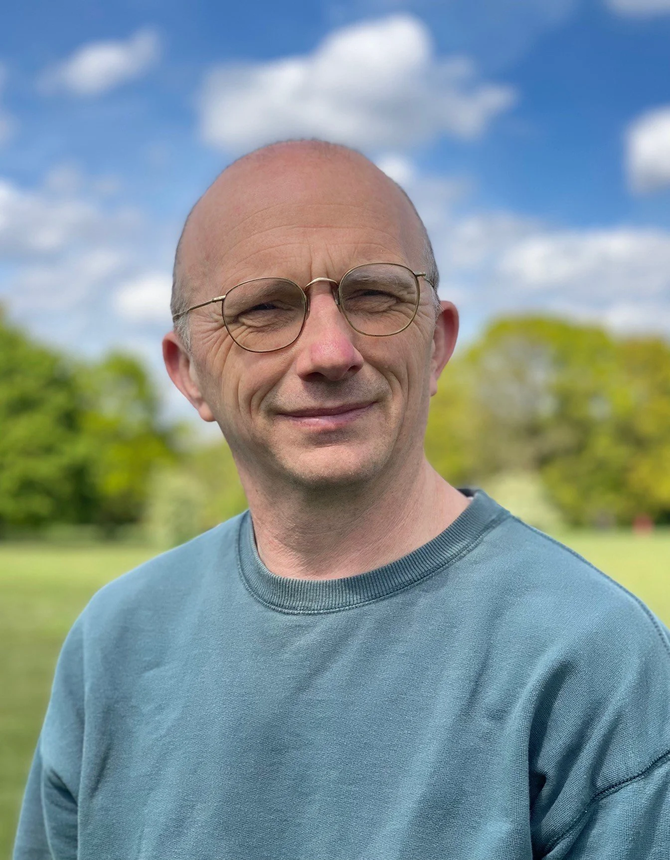 A middle-aged man with glasses and a bald head smiling outdoors on a sunny day, with trees and a blue sky with clouds in the background.