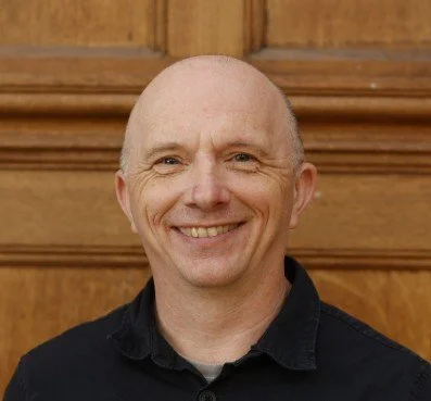 A smiling man with a shaved head, wearing a black shirt, standing in front of a wooden background.