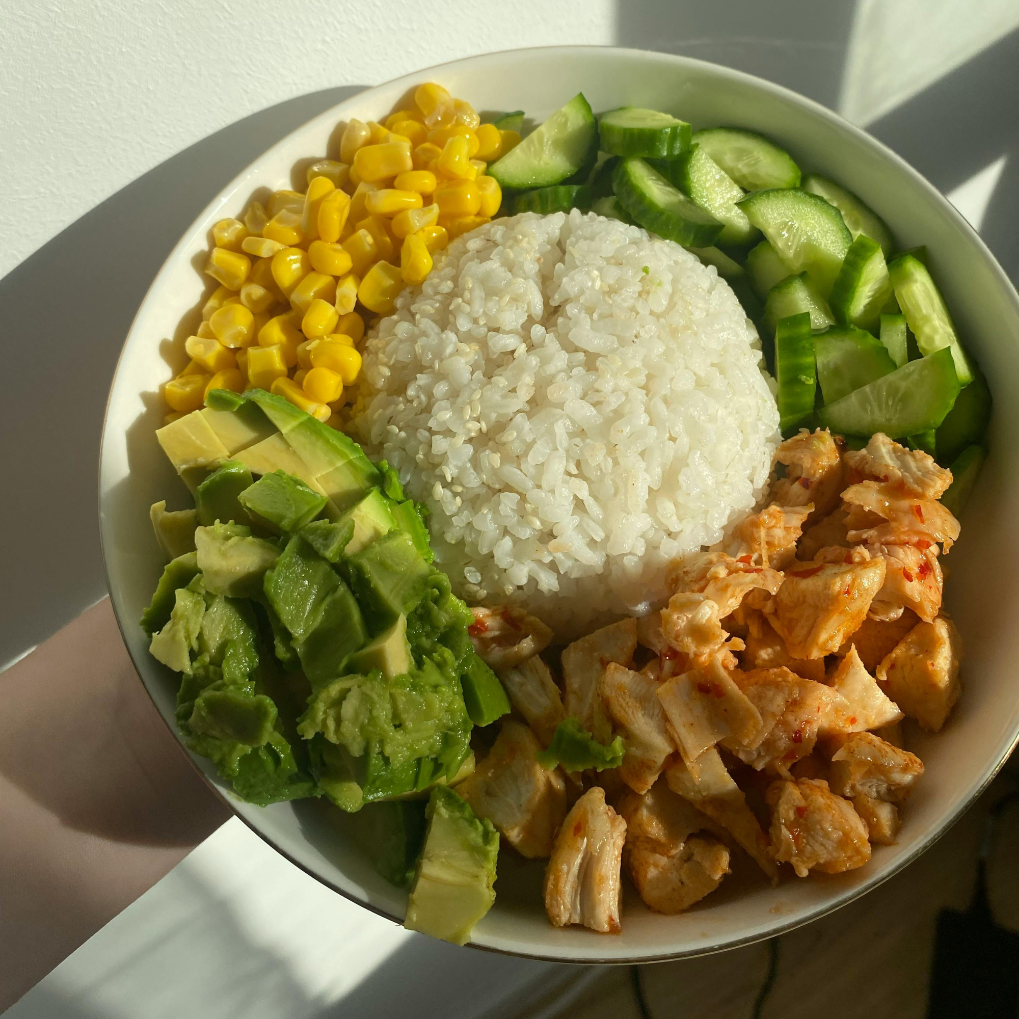 A bowl of rice with chopped chicken, sliced cucumbers, avocado, corn, and chopped green bell peppers.