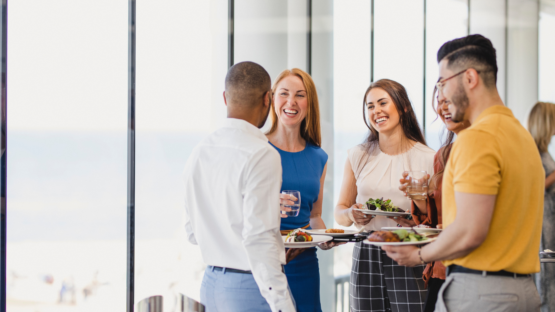 Group of five young professionals socializing with food and drinks in a modern office or event space with large windows