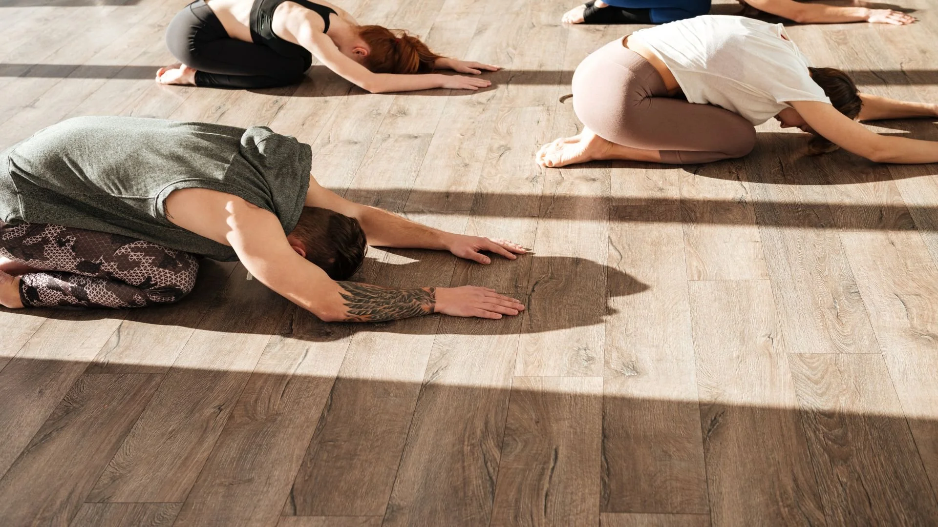 People participating in a yoga or meditation class, in Child's Pose on a wooden floor, with sunlight casting shadows.