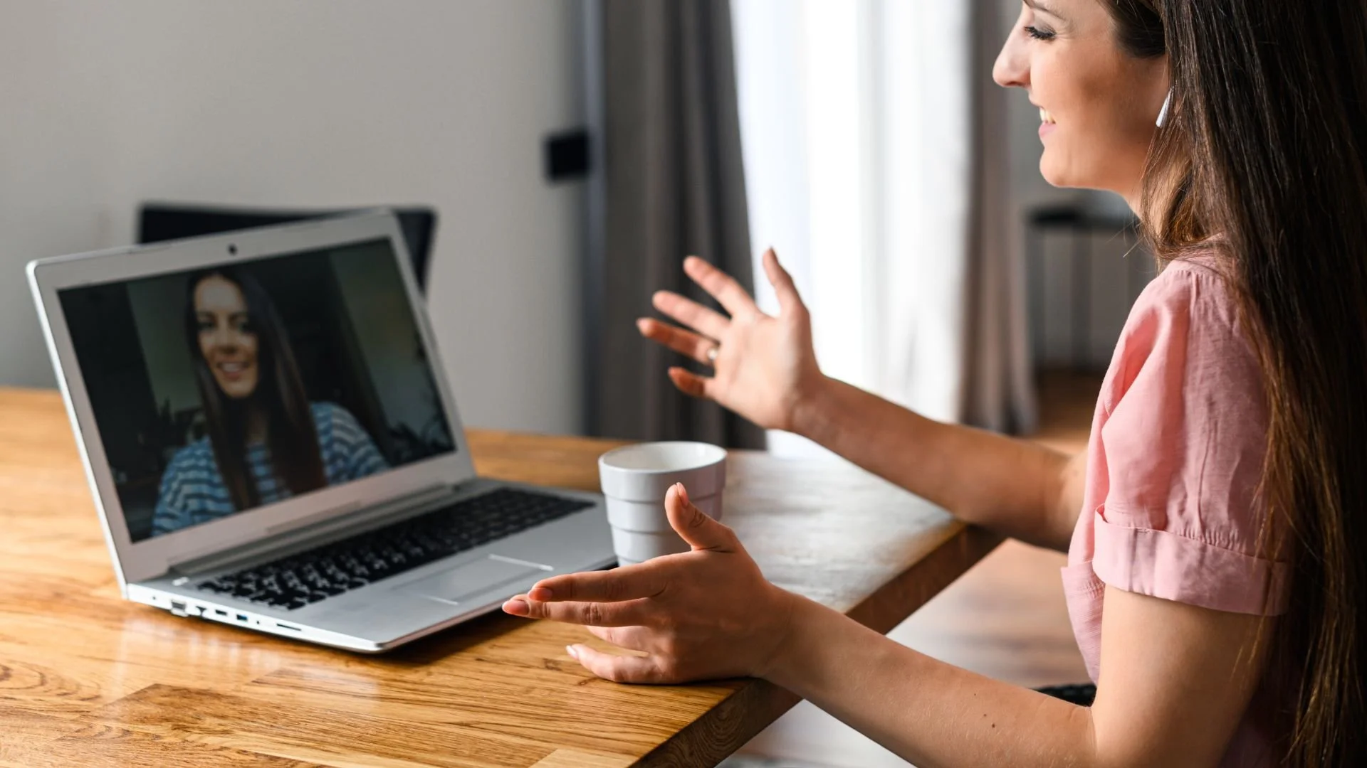 Woman with long brown hair wearing pink shirt participating in a video call on her laptop, waving at the screen in a brightly lit room.