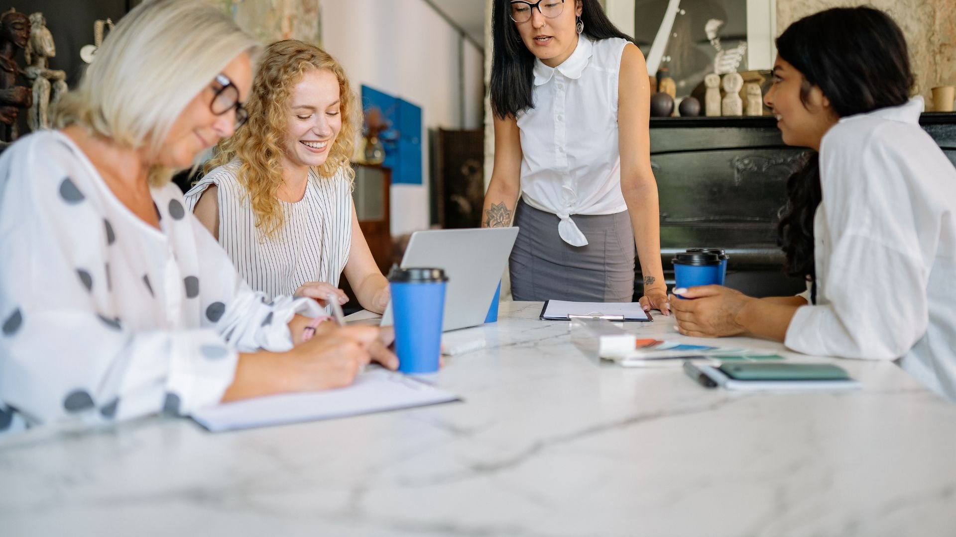 Four women working together at a table in a casual office setting, with notebooks, tablets, and coffee cups. One woman stands and discusses while others are engaged in writing or using devices.