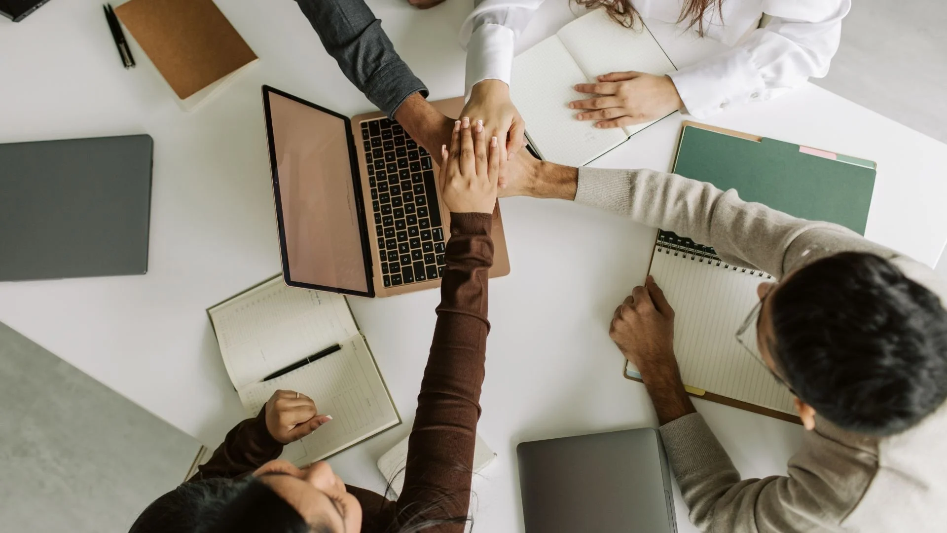 Four people at a white desk, reaching their hands together in a gesture of teamwork, surrounded by laptops, notebooks, and pens.