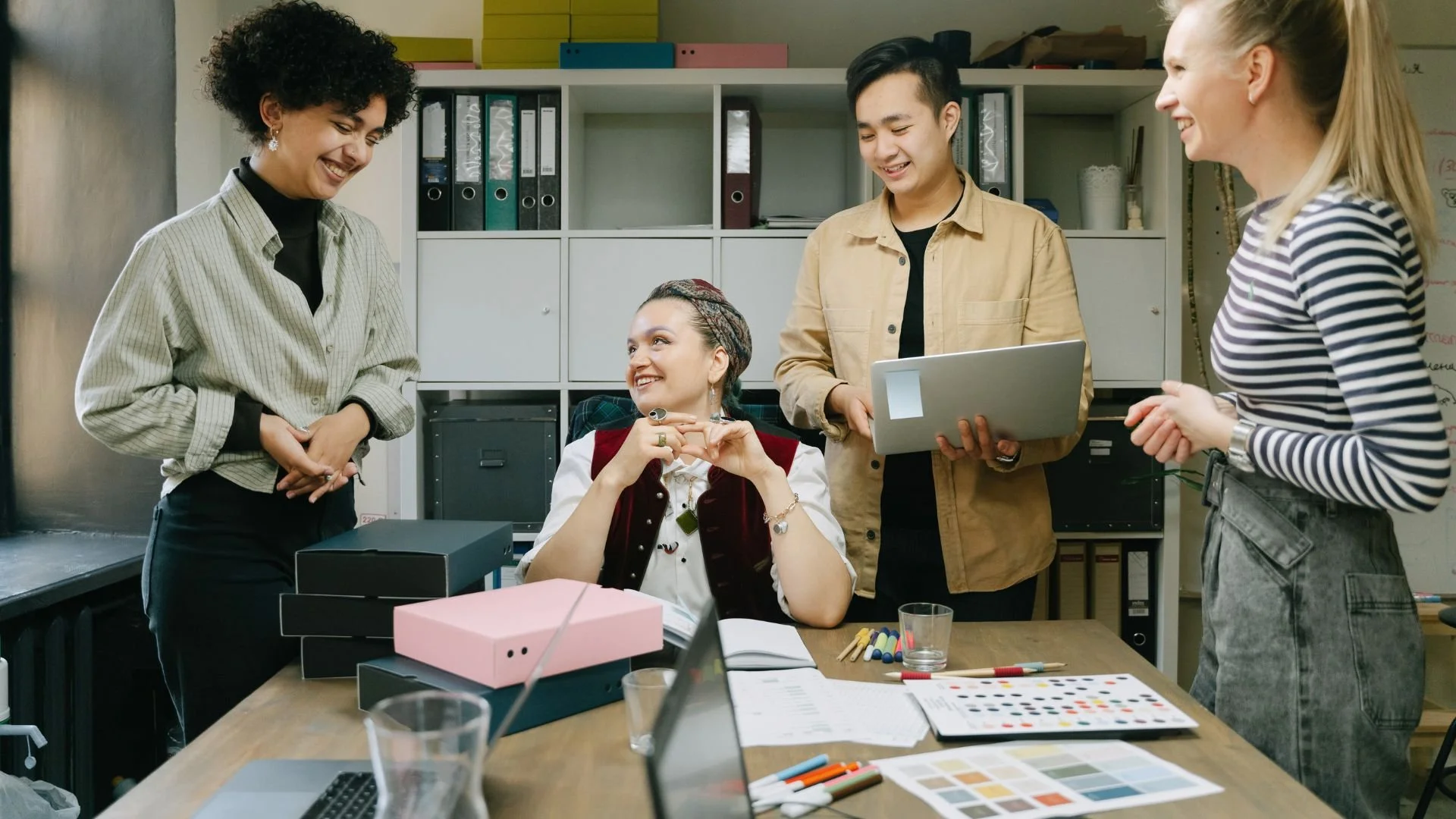 Group of five young professionals having a friendly discussion in an office, with a woman seated at the desk and four standing around, talking and smiling.