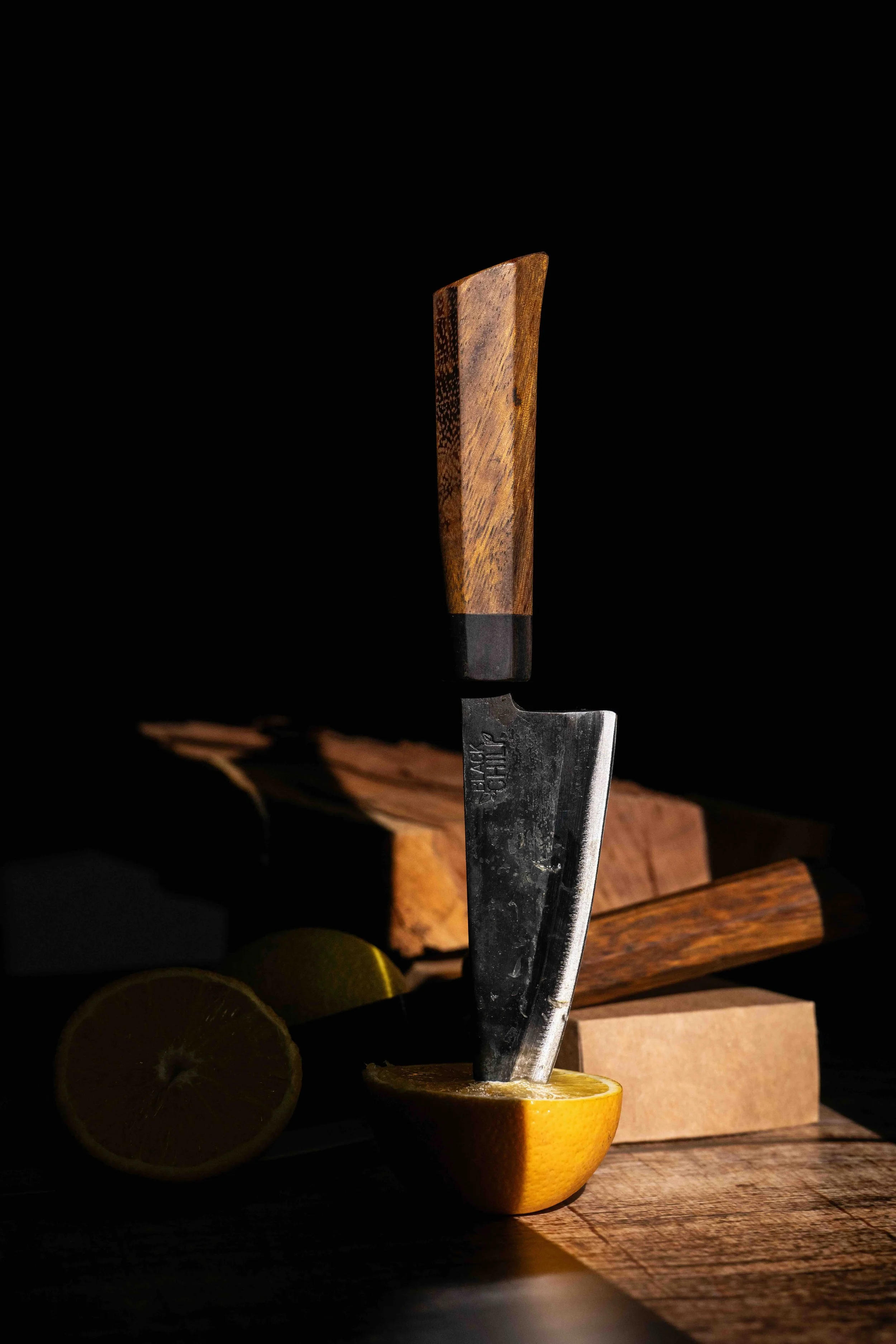 A knife with a wooden handle stands upright in a halved lemon on a wooden surface. There are additional lemon halves and wooden blocks in the background, with a dark backdrop and dramatic lighting.
