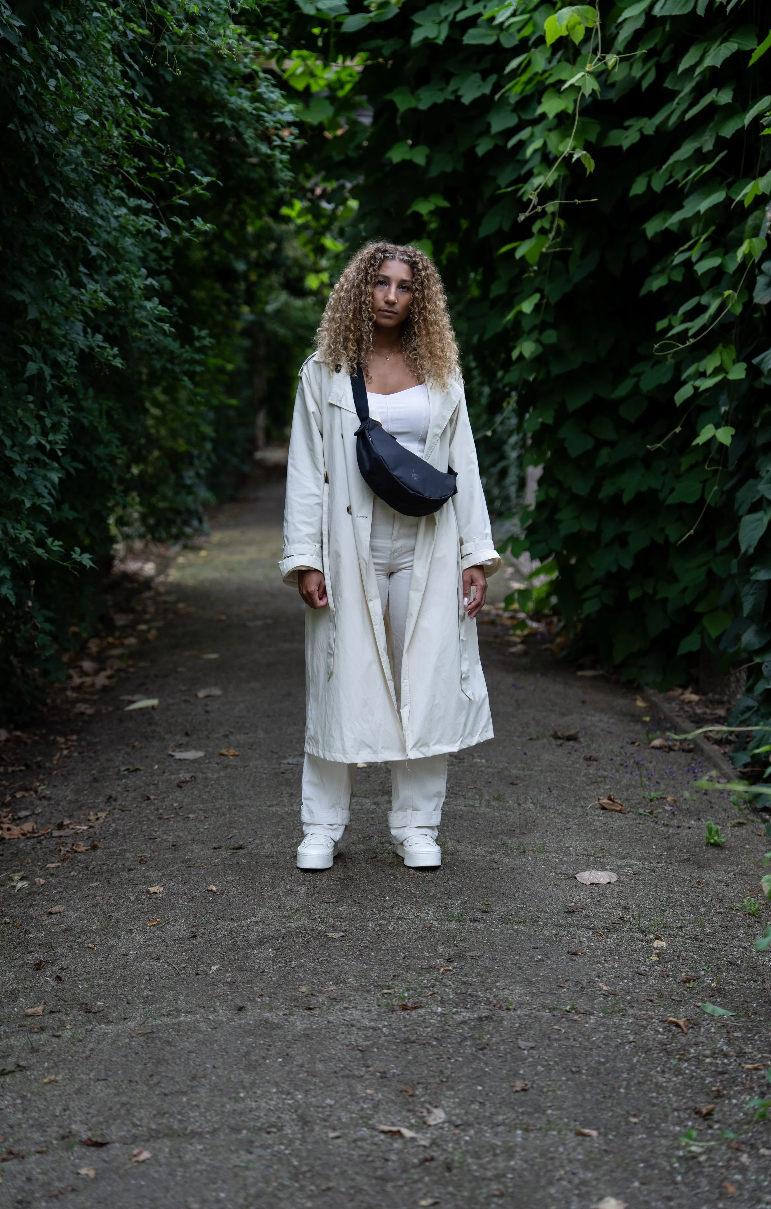 A woman with curly hair standing on a dirt pathway surrounded by green foliage, wearing a long beige trench coat, white pants, and a black crossbody bag.