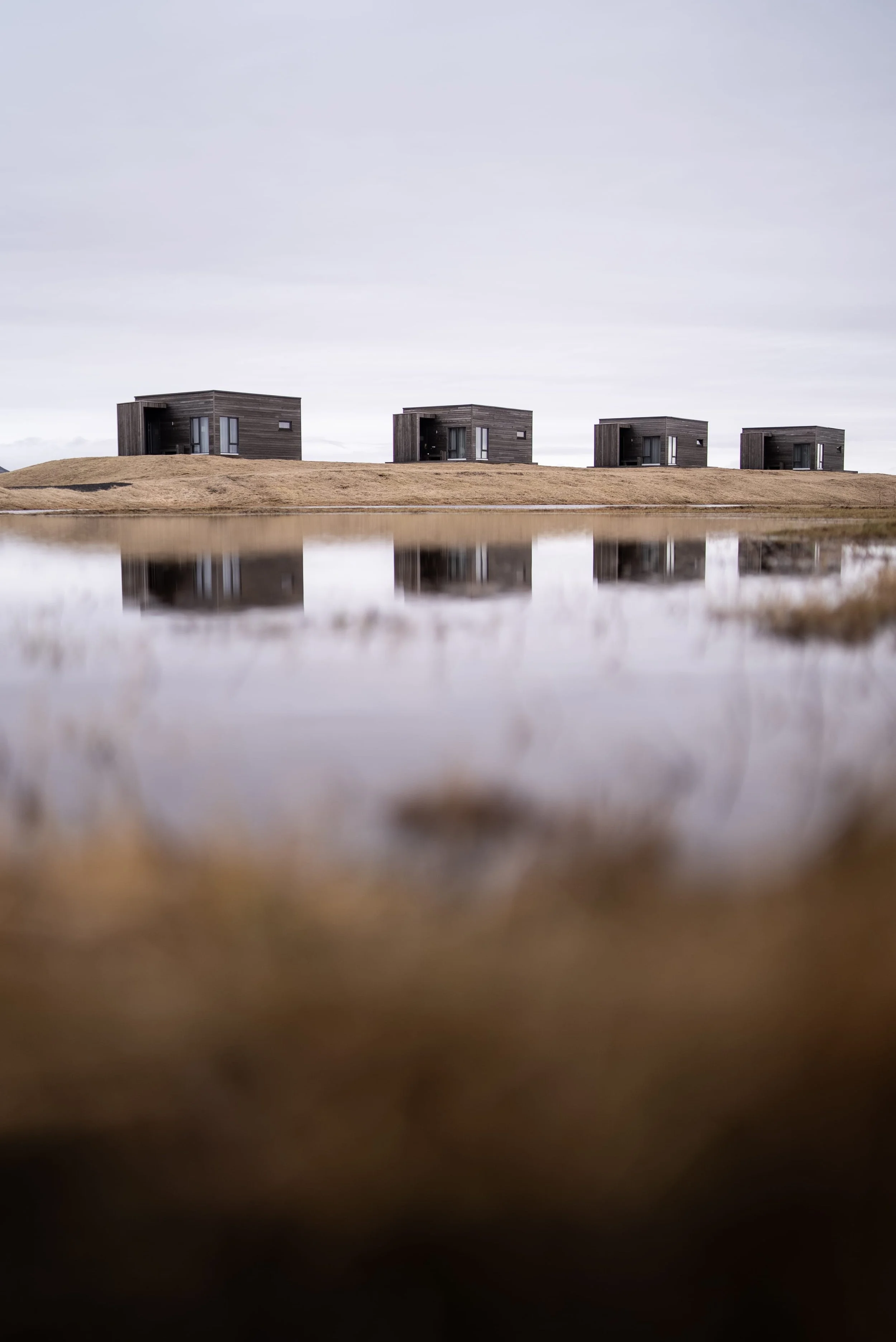 Four dark-colored modern houses on a grassy hill, reflected in a body of water in the foreground, under an overcast sky.