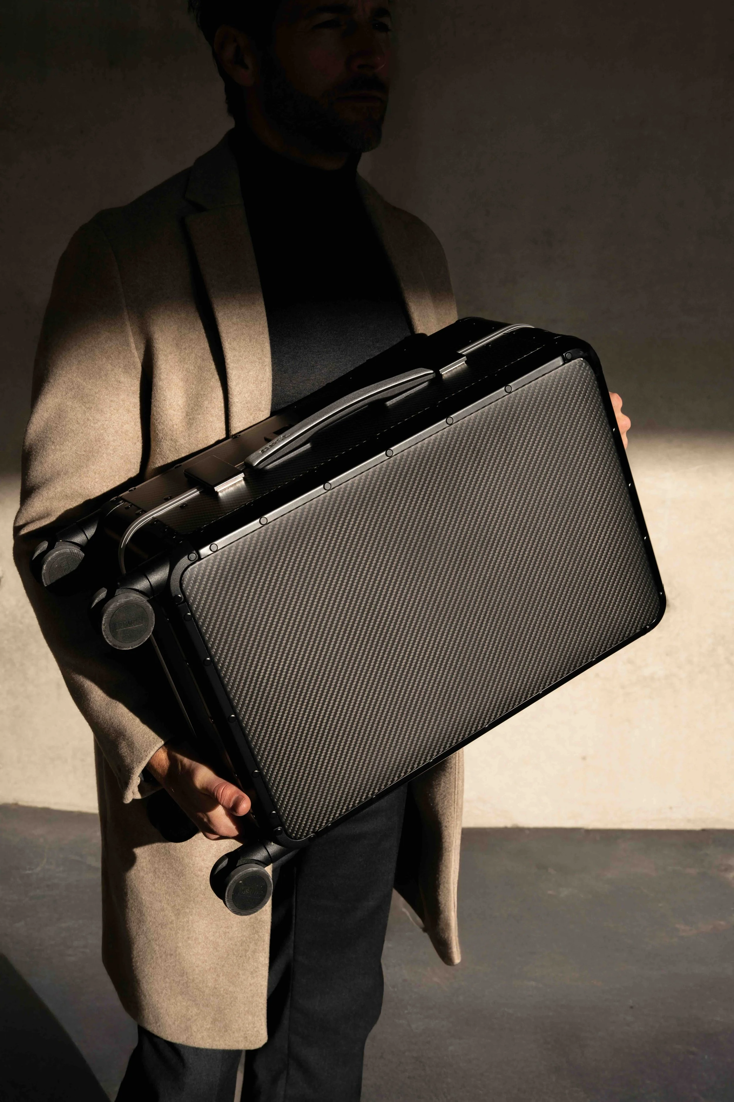 A man in a beige coat holding a large black briefcase with a textured surface, standing against a plain wall.