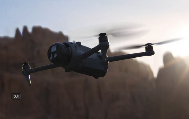 A DJI drone flying against a backdrop of mountains and a bright sky during sunset or sunrise.