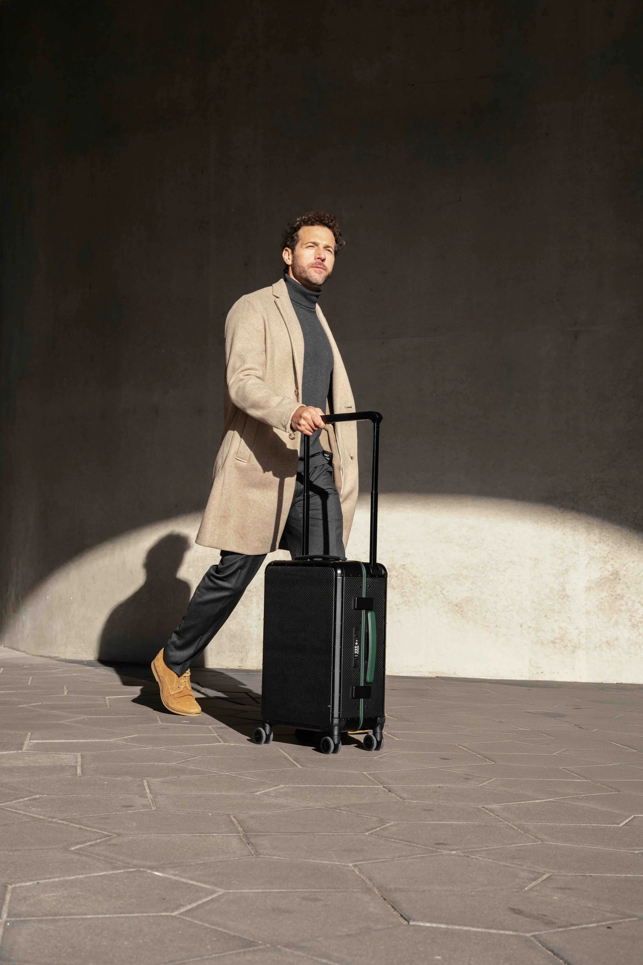 Man in beige coat, black turtleneck, black pants, and brown boots standing with a black rolling suitcase in sunlight.