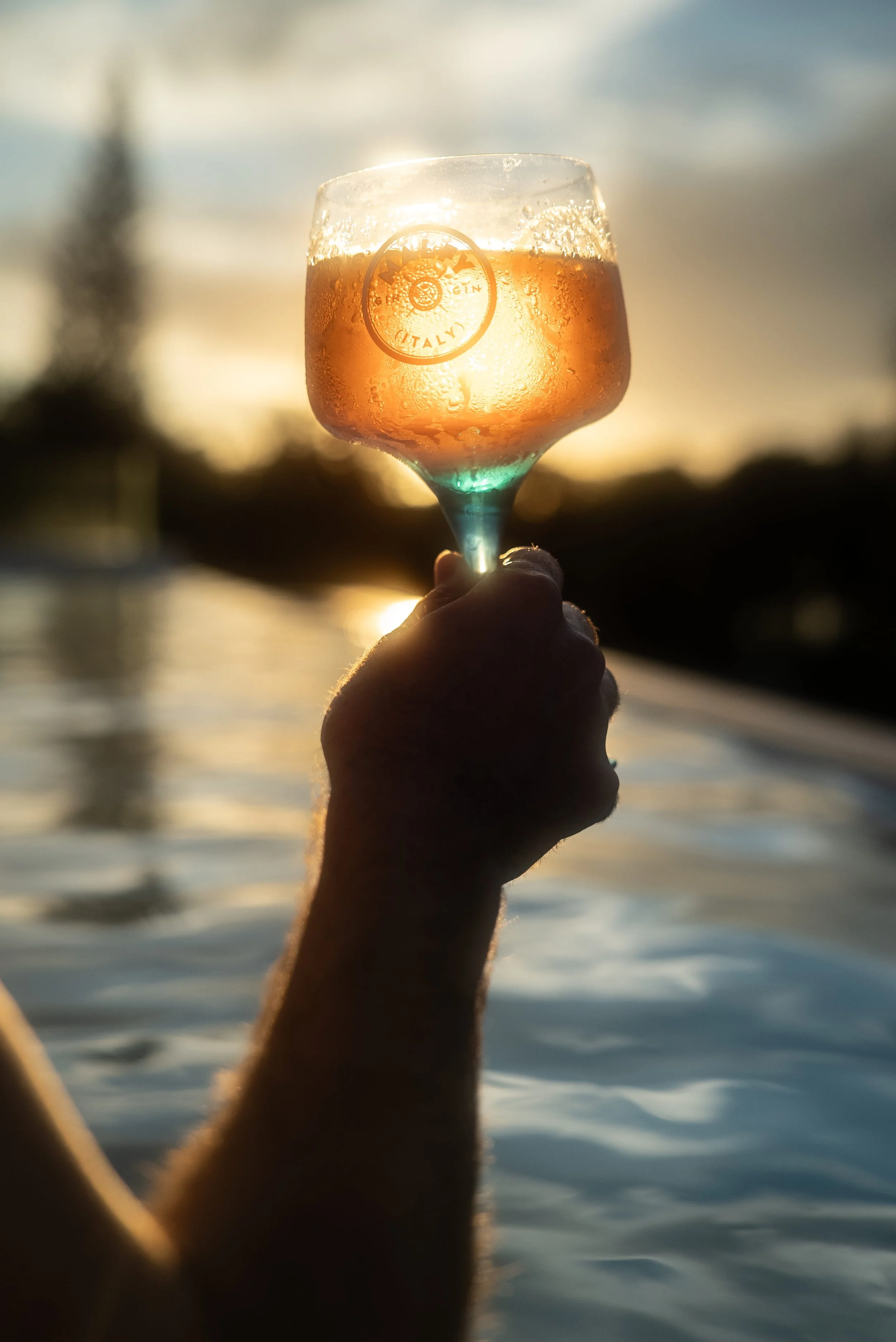 A hand holding a glass of rosé wine against a sunset background over water, with the glass condensation visible.