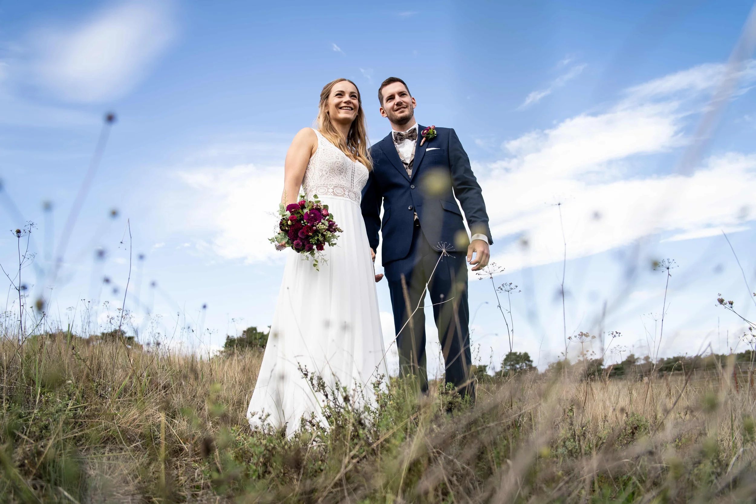 Bride and groom standing together outdoors on a grassy field, smiling and looking into the distance, with blue sky and clouds in the background.