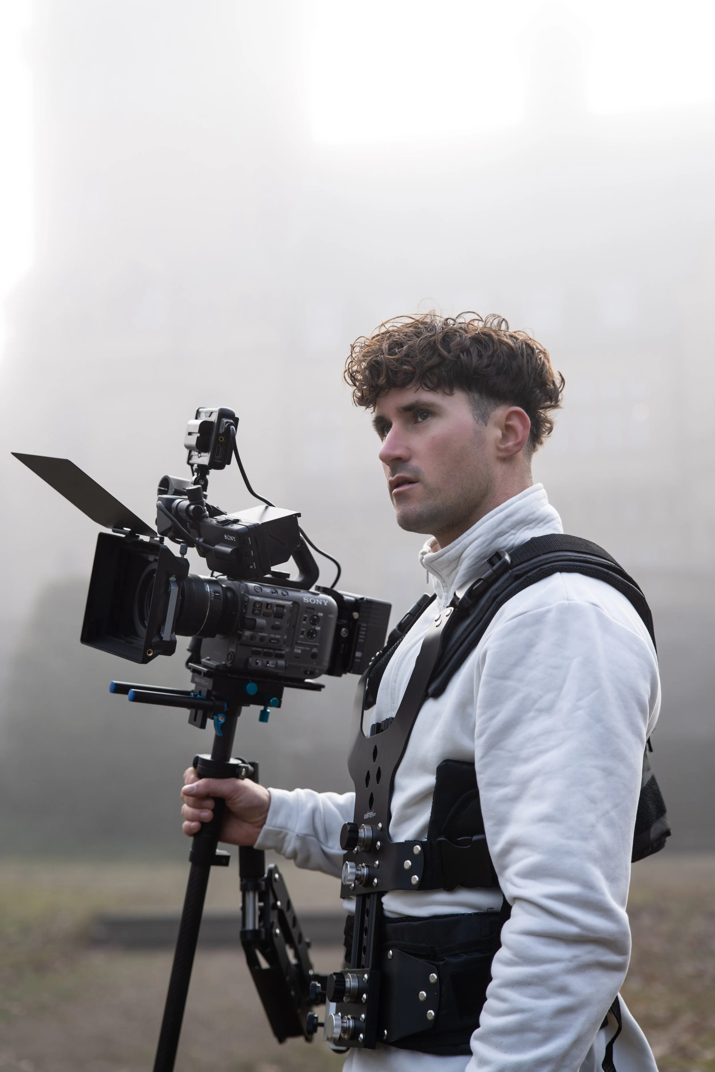 Male filmmaker with camera and stabilizer gear outdoors in a foggy or overcast setting.