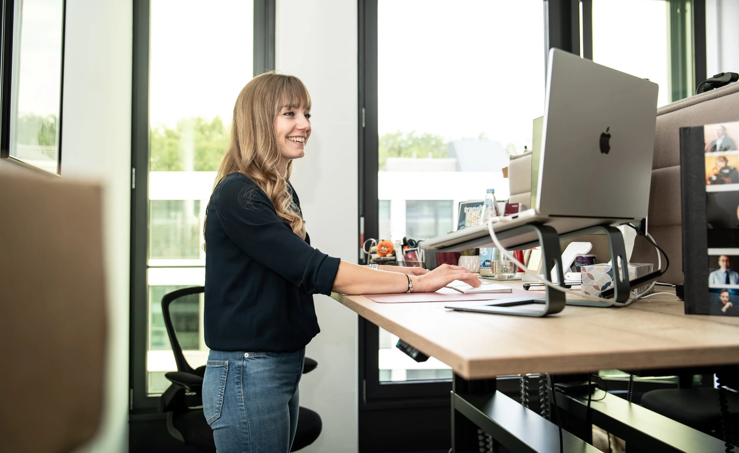Woman with long wavy hair working at a standing desk in an office, smiling, with a computer and various office supplies.