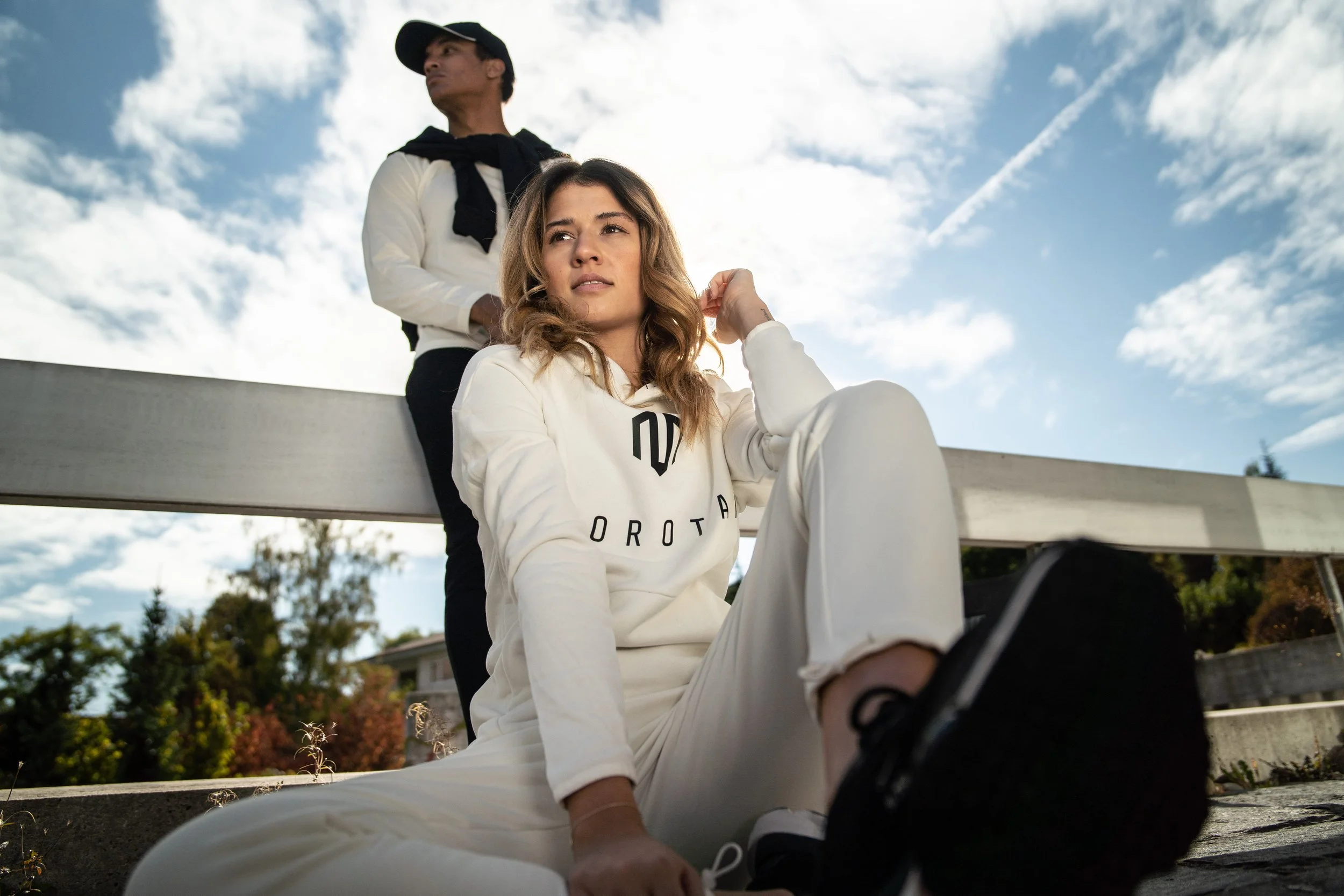 Two young adults, a woman sitting on a ledge and a man standing behind her, outdoors on a sunny day with a partly cloudy sky. The woman has blonde hair and is wearing a white hoodie and pants, while the man has black hair and is dressed in a white shirt with a black sweater tied around his shoulders.