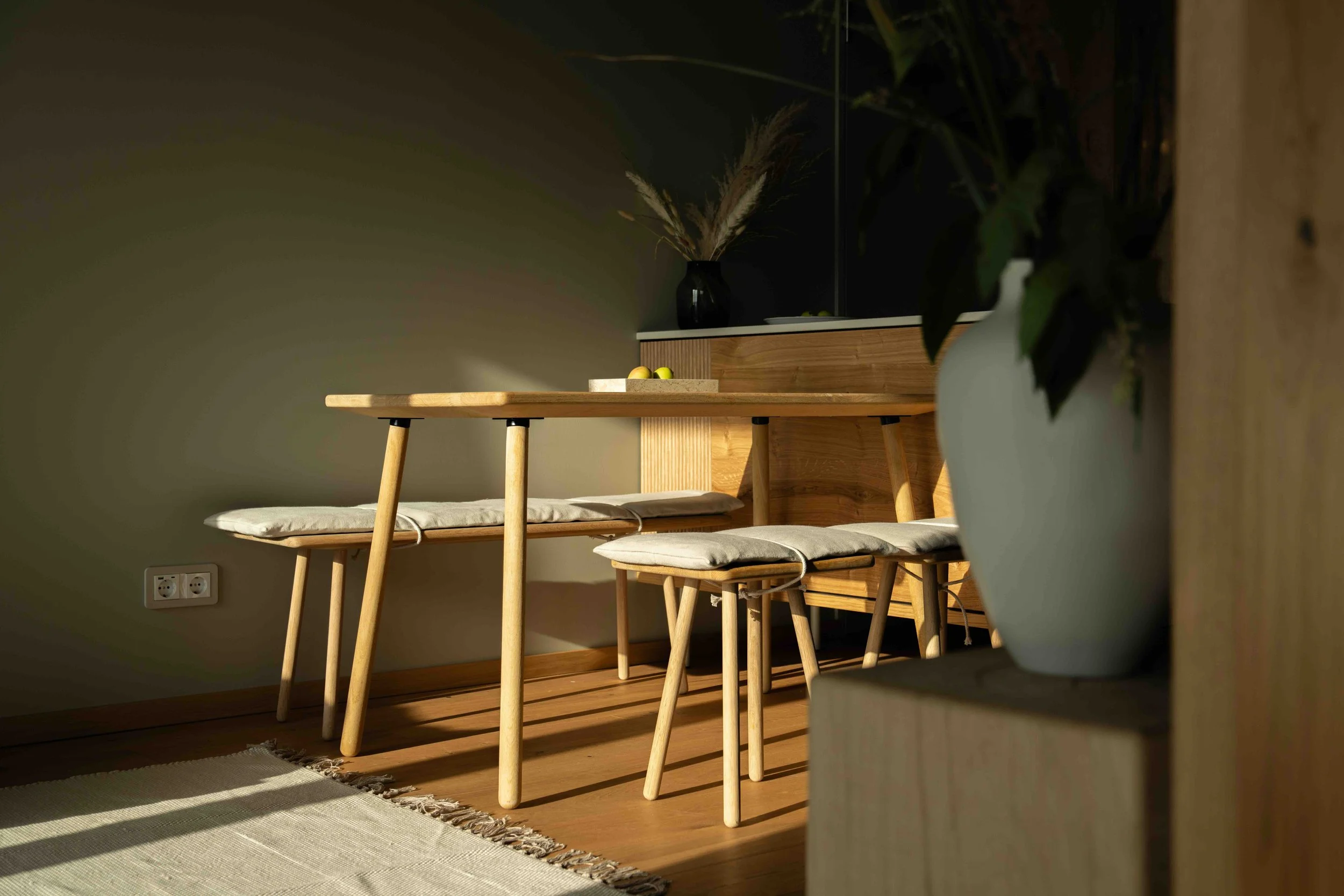 Dining area with wooden table, two matching wooden benches with cushions, a large white vase with greenery, and a wooden sideboard with a black vase and dried plants, illuminated by warm lighting.
