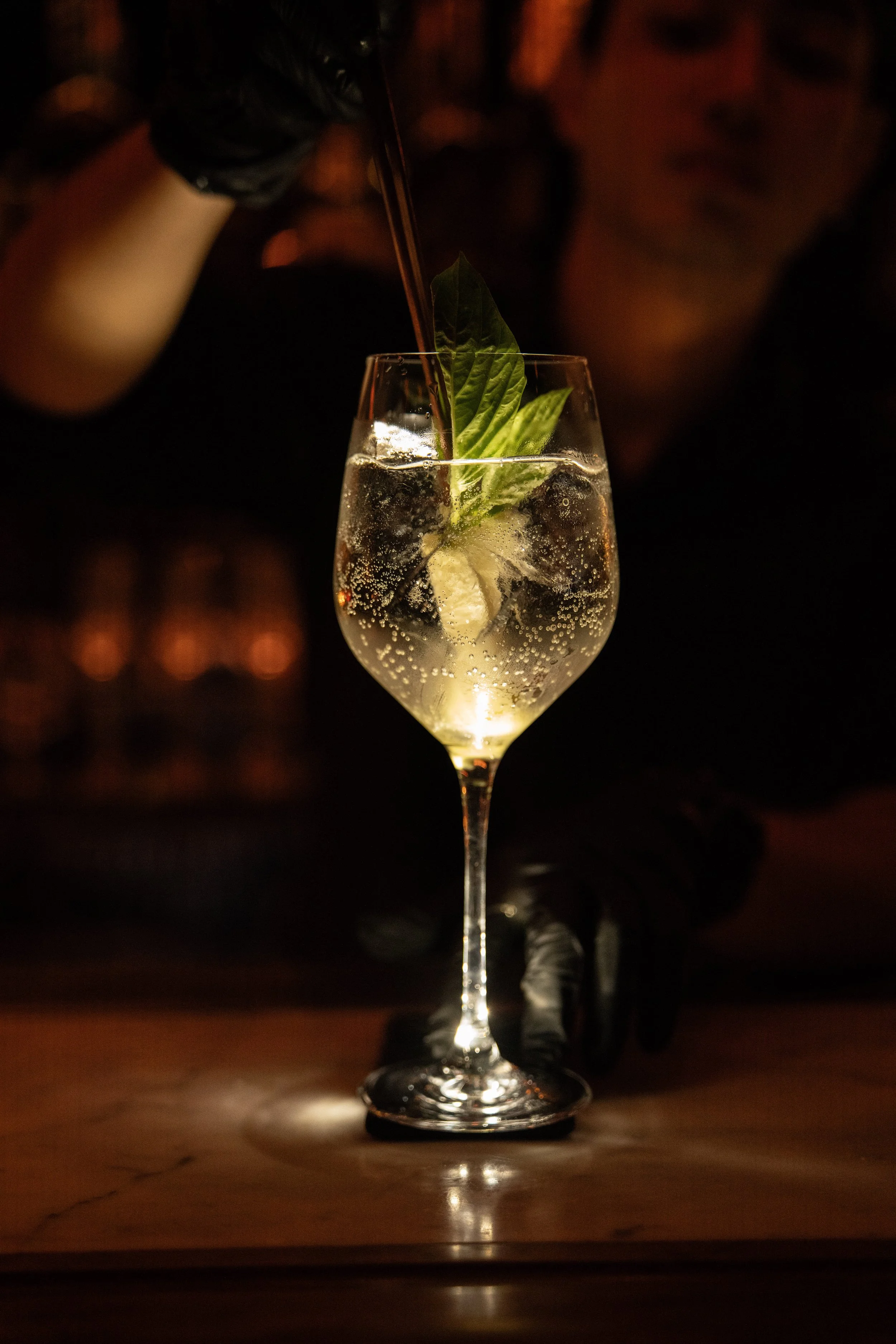 A bartender garnishing a tall wine glass filled with a clear fizzy beverage, ice, fresh mint, and a lemon wedge with a straw, against a dark background.