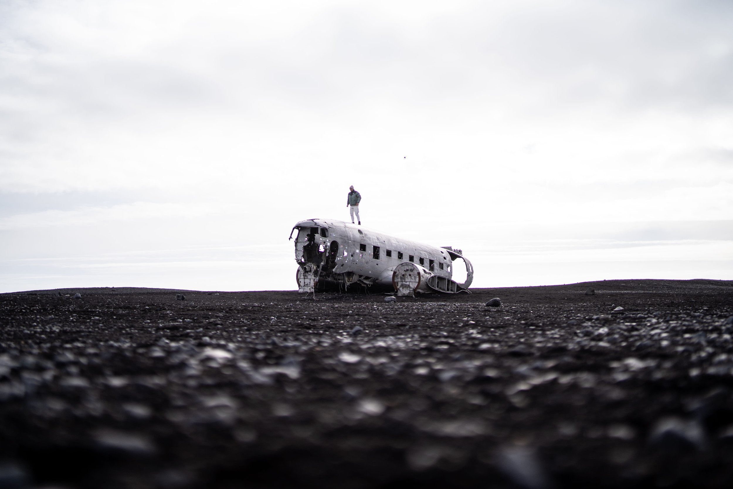 A person standing on top of a wrecked airplane fuselage in a barren, rocky landscape under a cloudy sky.