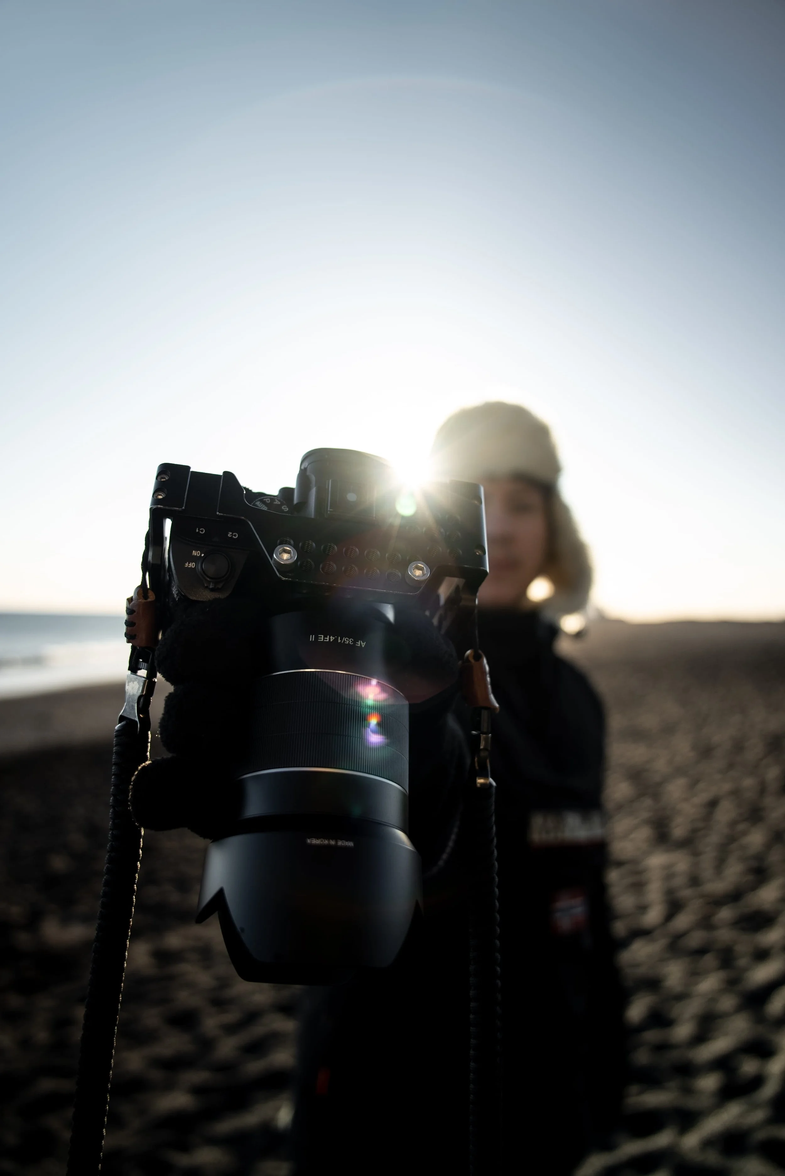 Person holding a camera pointed towards the camera with the sun shining behind them on a beach.
