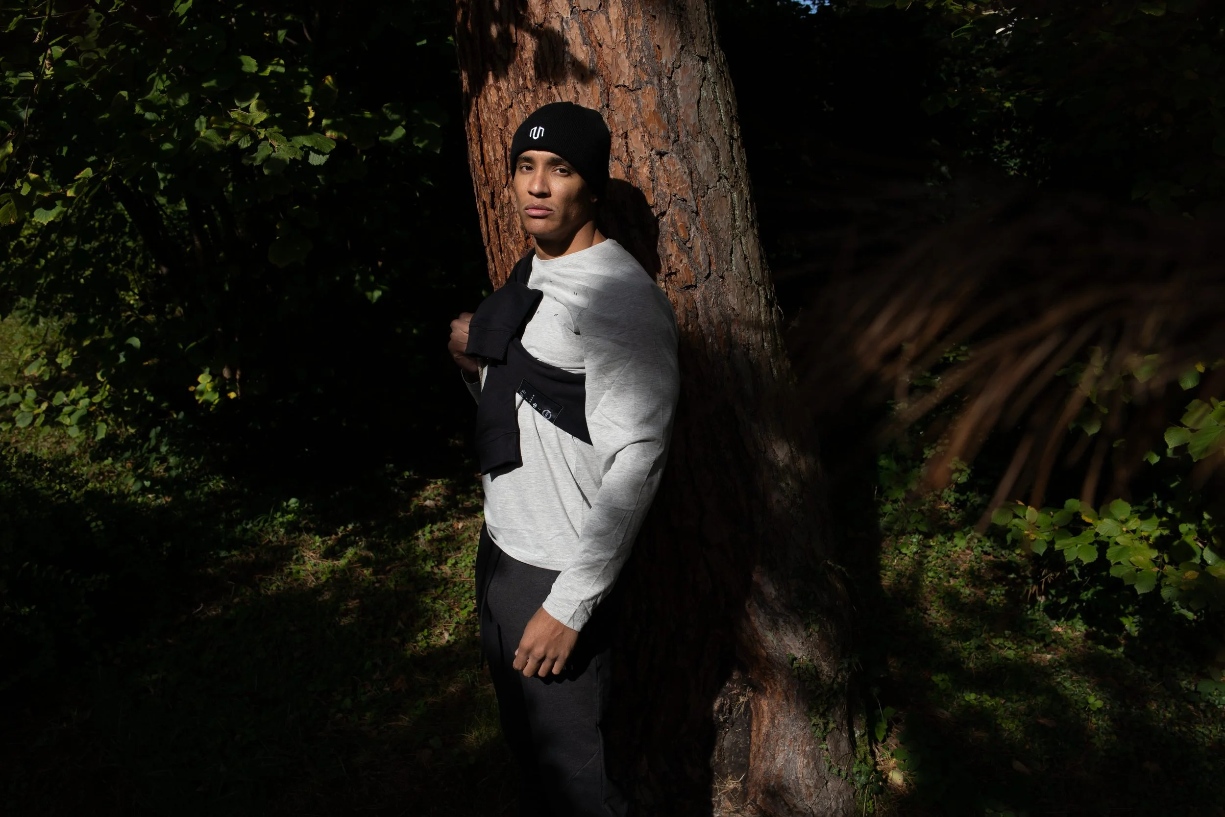 A young man leaning against a tree in a wooded area, wearing a black beanie, a light gray sweatshirt, and black pants, holding a black jacket over his shoulder.