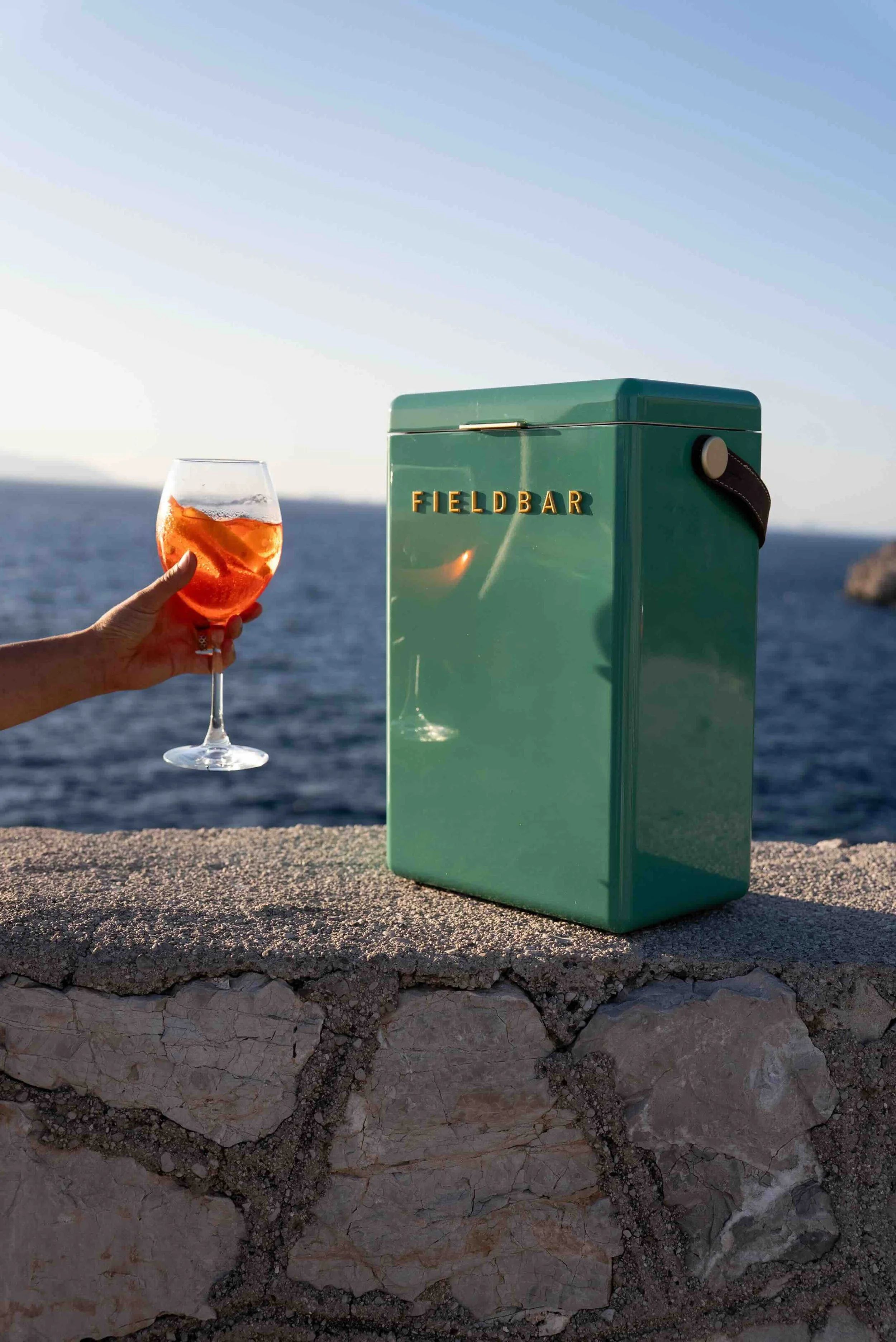 A hand holding a glass of rose-colored drink with ice cubes, placed on a stone wall by the sea, with a teal-colored box labeled 'FIELD BAR' on the wall.