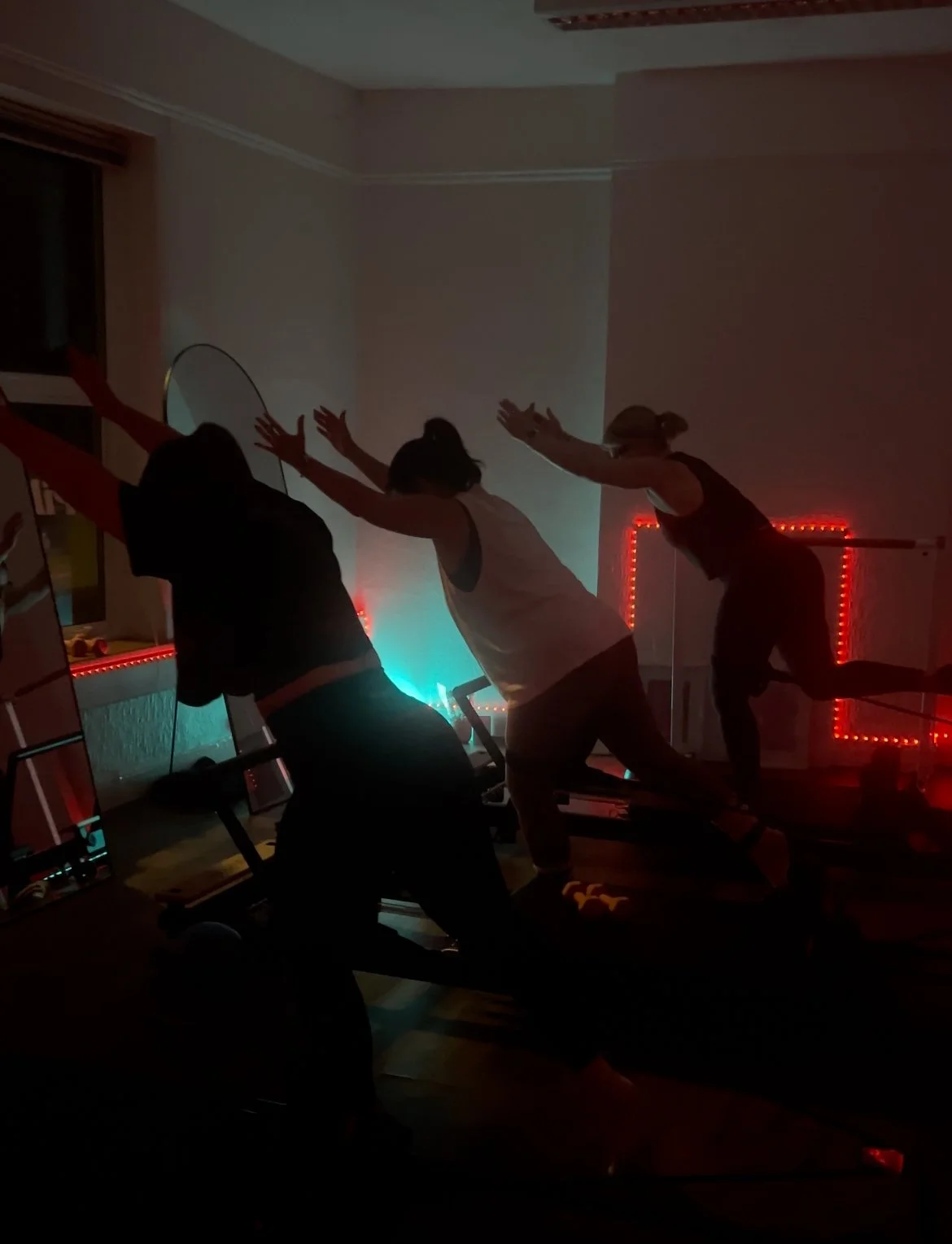 Three women on exercise bikes stretching with arms outstretched in a dimly lit room with red LED lights in the background.