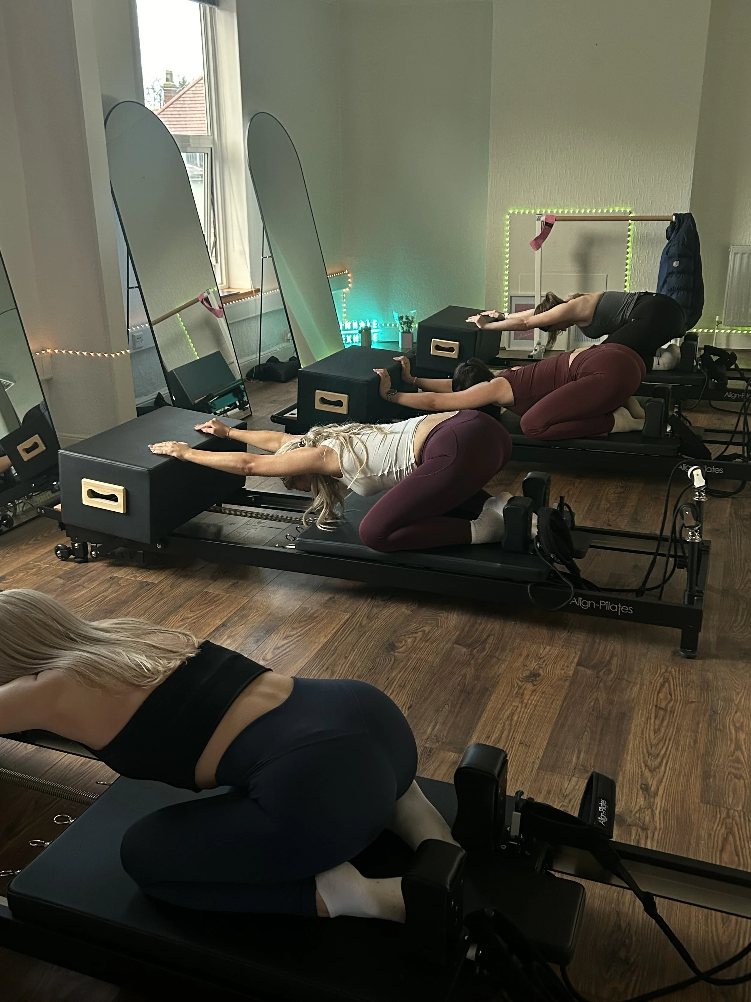 Three women doing Pilates on reformer machines in a room with wooden floors, large mirrors, and windows. Two women are on the reformers, and one woman is on a mat in the foreground.