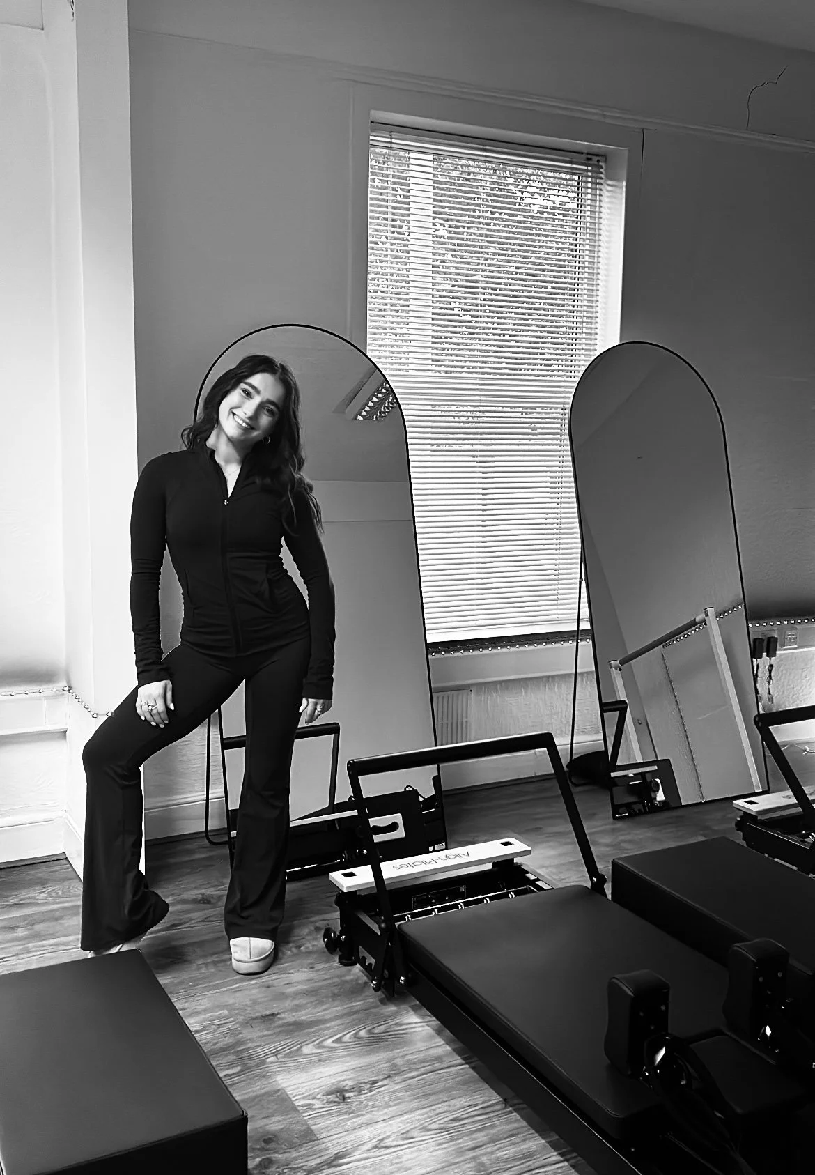 A woman smiling and posing in a room with mirrors and fitness equipment, in black and white.