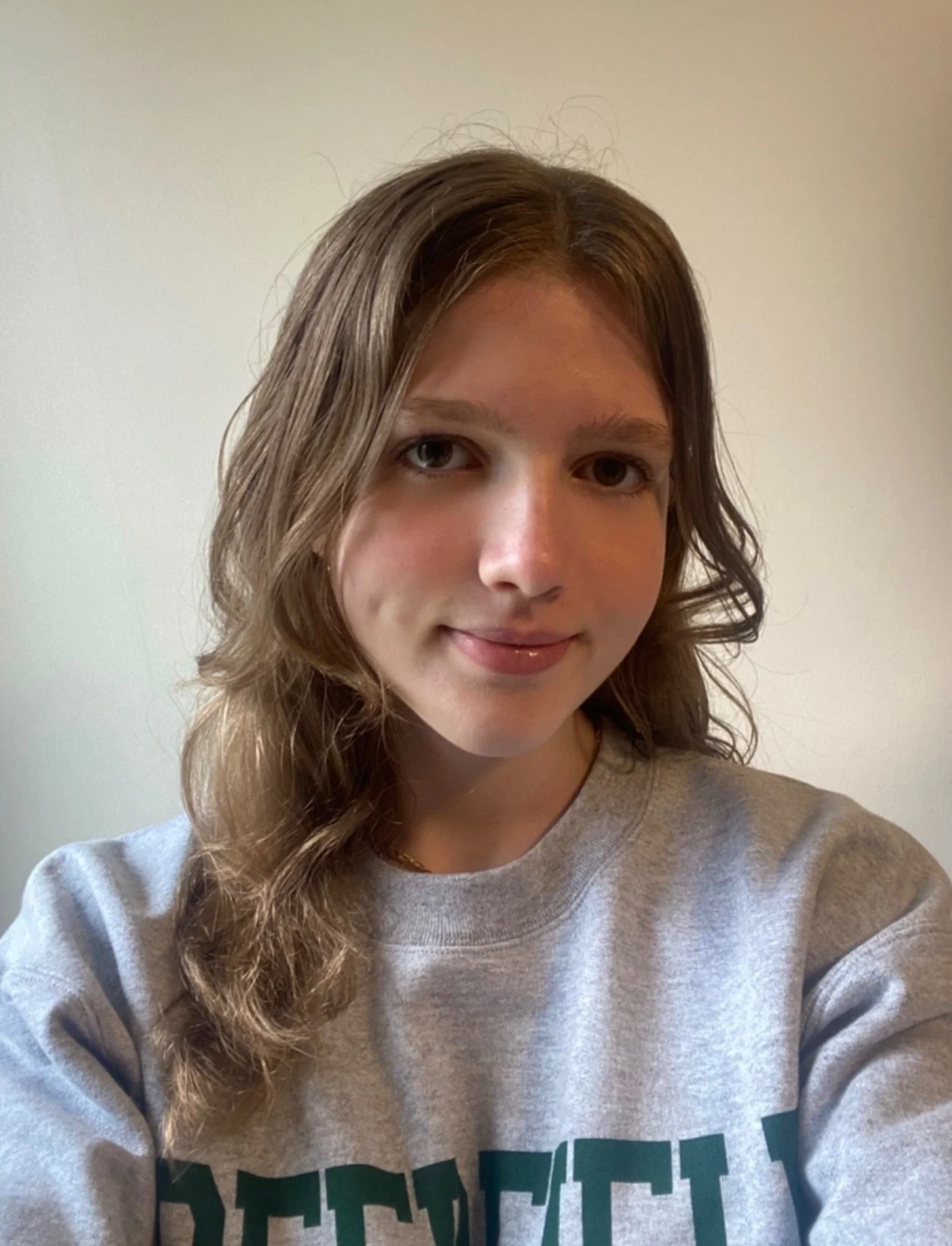 A high school student filmmaker with long wavy hair, wearing a gray sweatshirt, smiling softly against a plain light-colored wall.