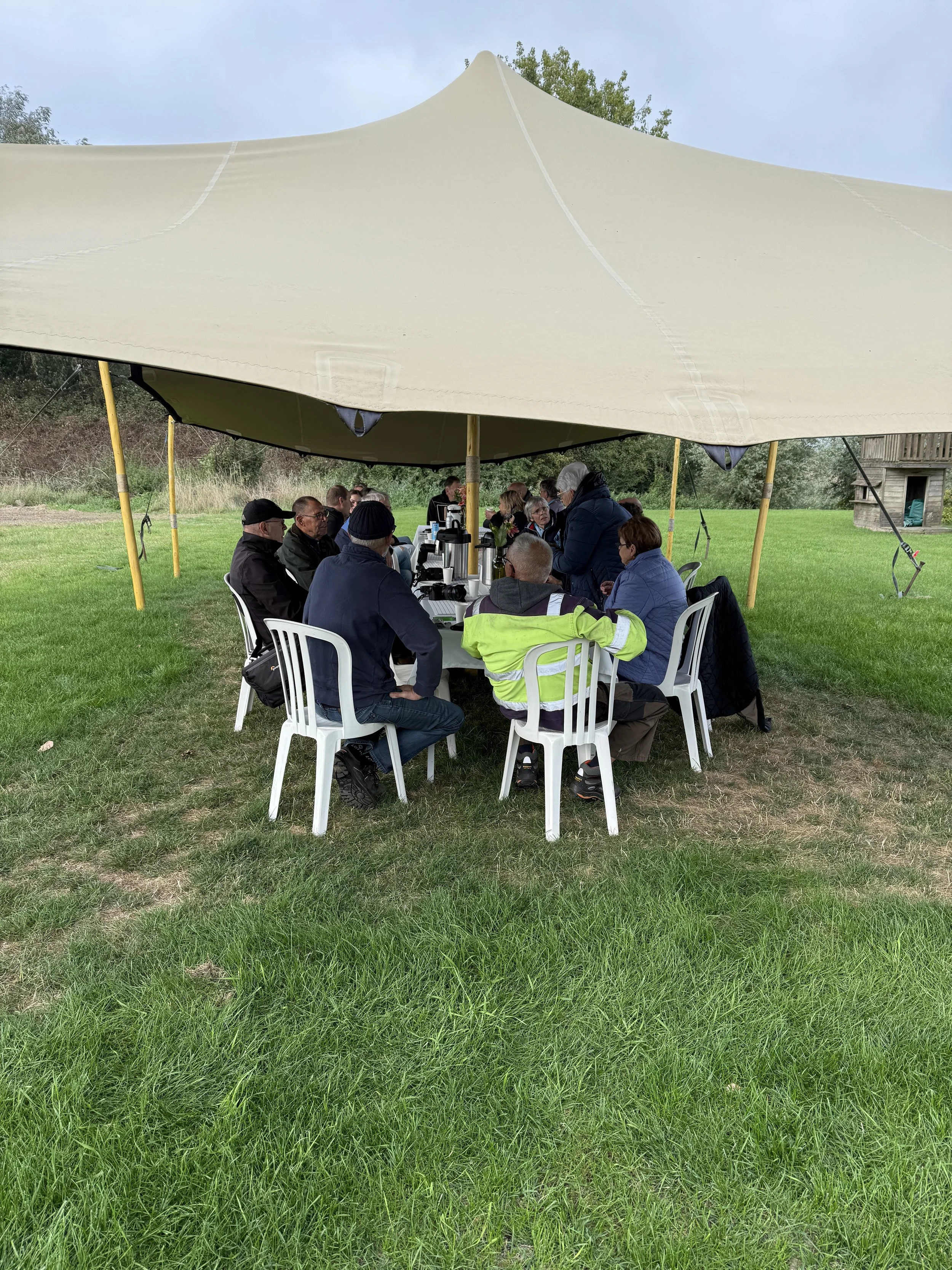 Een groep mensen zit onder een grote tent op een grasveld, tijdens de plekdagen op wijngaard bossenblie in Zeeuws-Vlaanderen