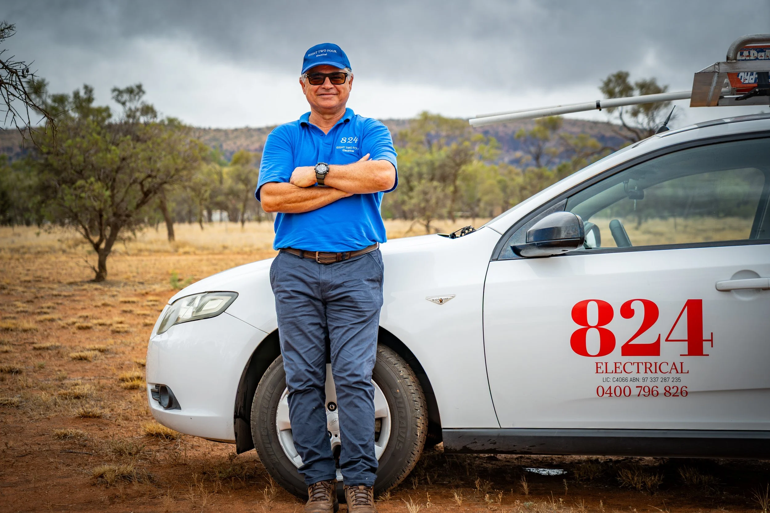 A man wearing sunglasses and a blue polo shirt with the logo '824 Electrical' stands with crossed arms next to a white electrical service vehicle on a dirt field with trees and cloudy sky in the background.