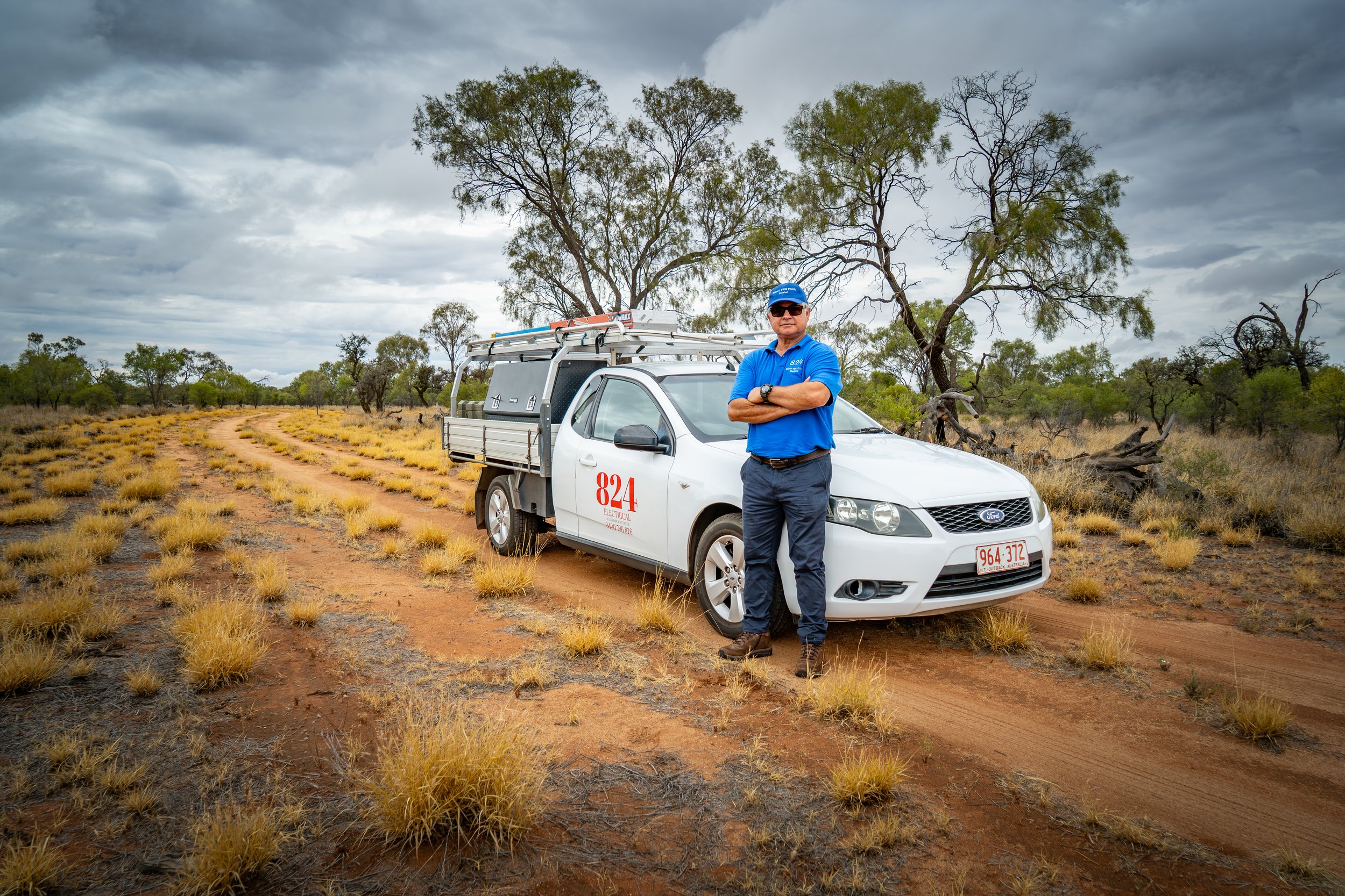A man in a blue uniform and cap standing with arms crossed next to a white utility truck on a dirt path in a semi-arid landscape with sparse trees and yellow grass under a cloudy sky.