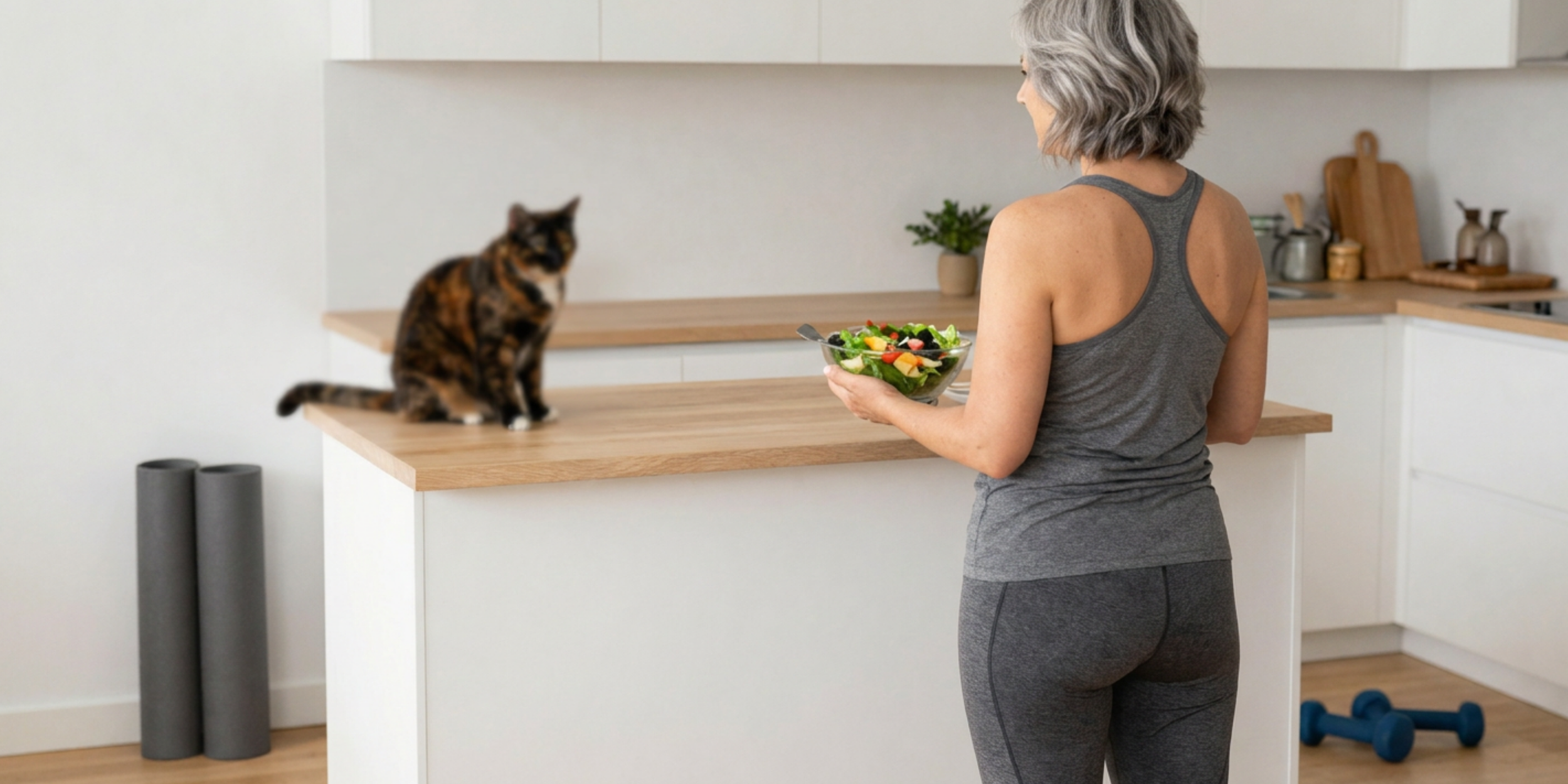 Une femme avec des cheveux gris portant un débardeur gris et un legging gris prépare une salade dans une cuisine moderne, devant un chat assis sur le comptoir.