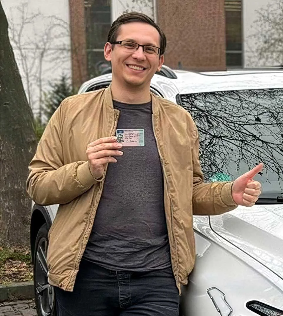 Ein Mann steht vor einem Auto, hält einen Führerschein in der Hand und zeigt mit dem Daumen nach oben. Es ist draußen, und im Hintergrund sind Gebäudefassaden und Bäume sichtbar.