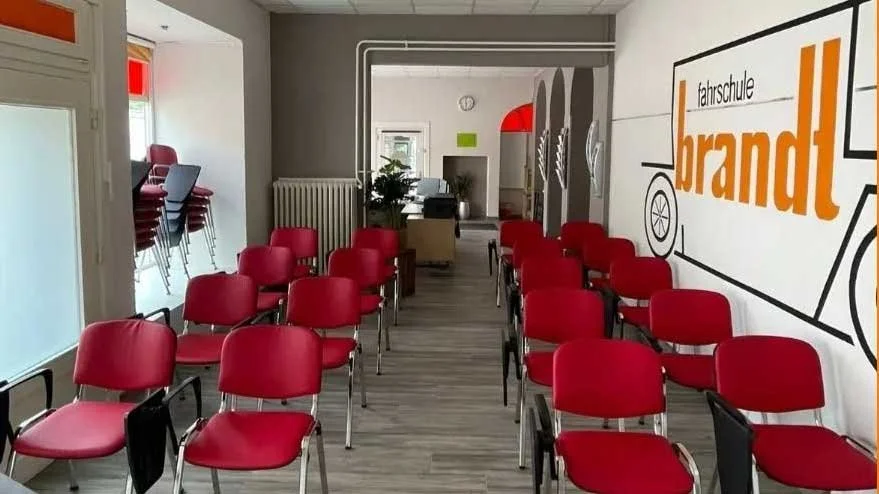 Ein Schulungsraum mit roten Stühlen, einem Fenster und einer großen Wand mit einem Logo der Fahrschule Brandt.