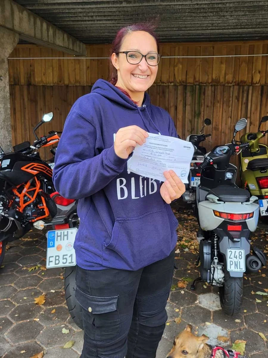 Eine Frau mit Brille und lila Hoodie hält eine Quittung, umgeben von Motorrädern und einem Hund, draußen auf einem gepflasterten Bereich bei bewölktem Wetter.