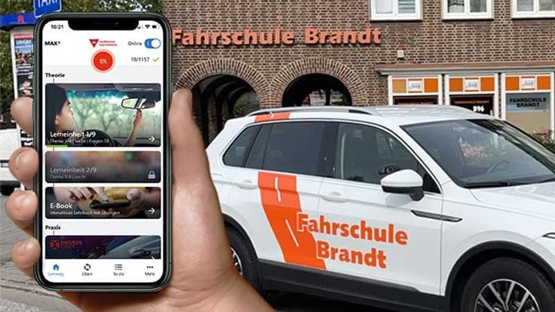 Eine Person hält ein Smartphone vor ein Auto, das vor einer Fahrschule namens 'Fahrschule Brandt' parkiert ist. Das Smartphone zeigt eine Lern-App für den Führerschein.