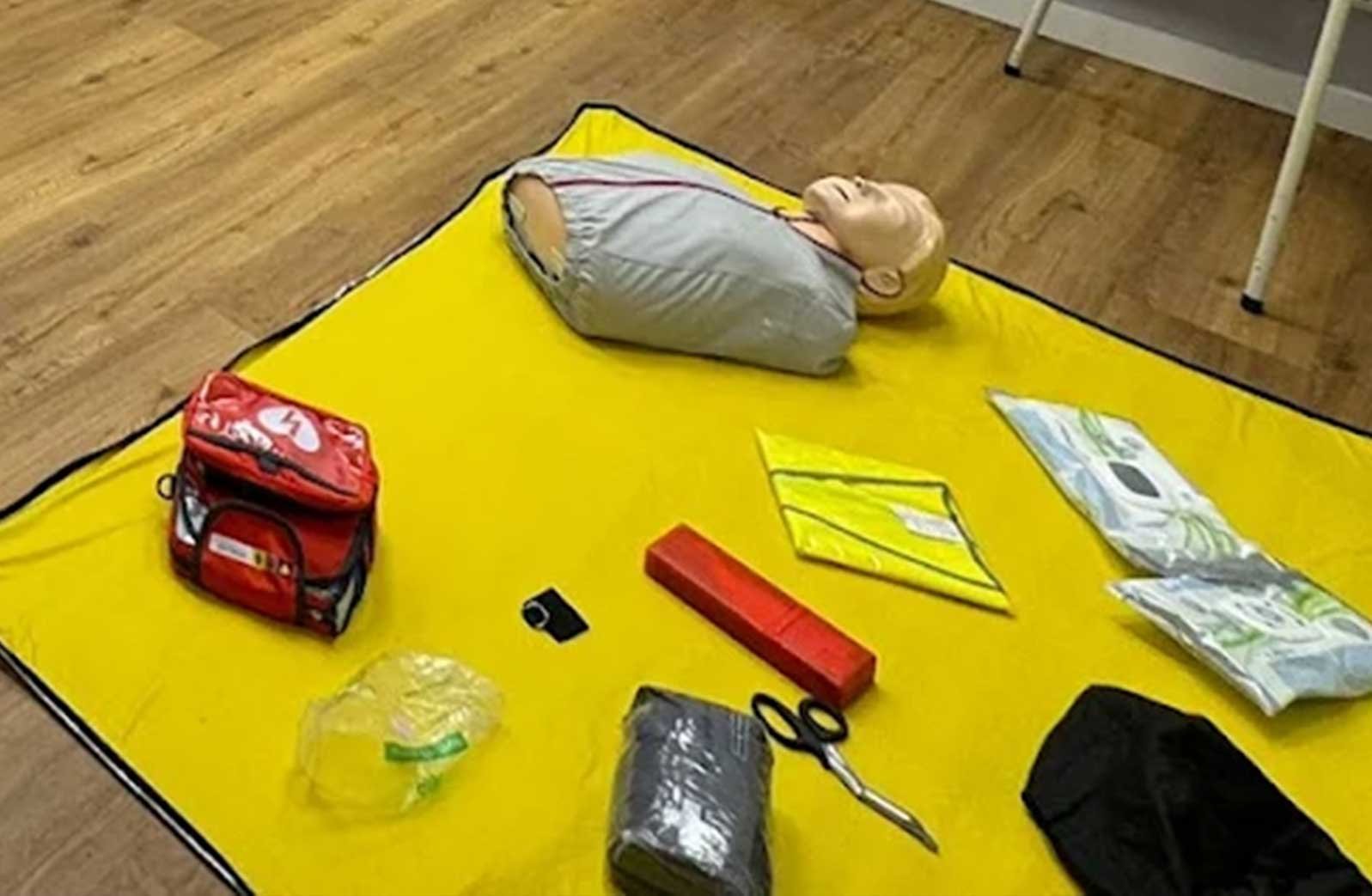 Blick auf einen Notfallrucksack, einen Cuttermesser, ein rotes Kissen, einen schwarzen Gegenstand, ein Paar Scheren, eine gelbe Mappe, eine Packung Feuchttücher, ein abgepacktes Sandwich, einen Beutel mit Sandwich, eine blaue Packung mit Blättern, eine Person in einem Schlafsack liegt auf einem gelben Tuch auf dem Boden.