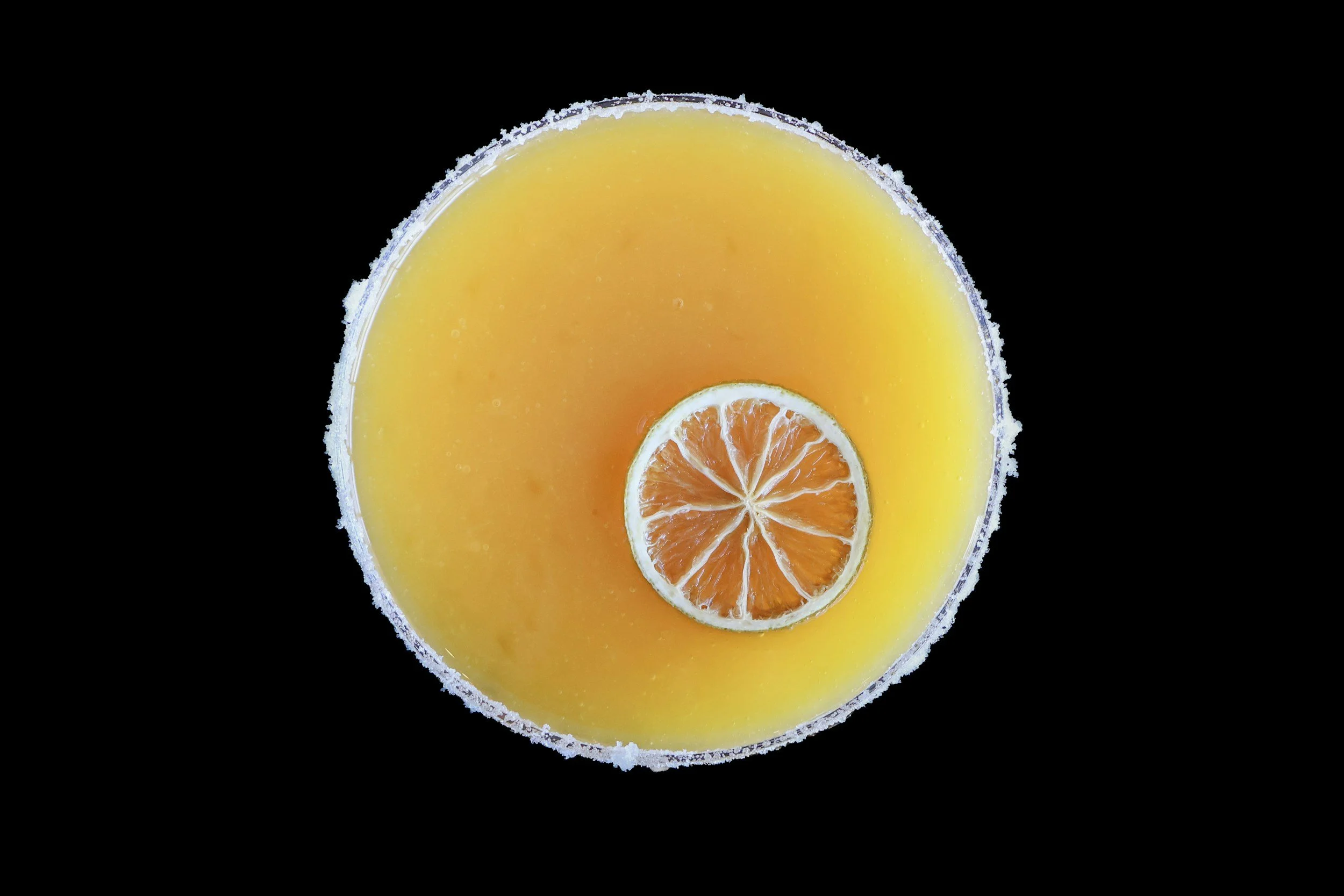 Top view of a yellow-orange beverage with a salted rim and a slice of lemon float on top, against a black background.