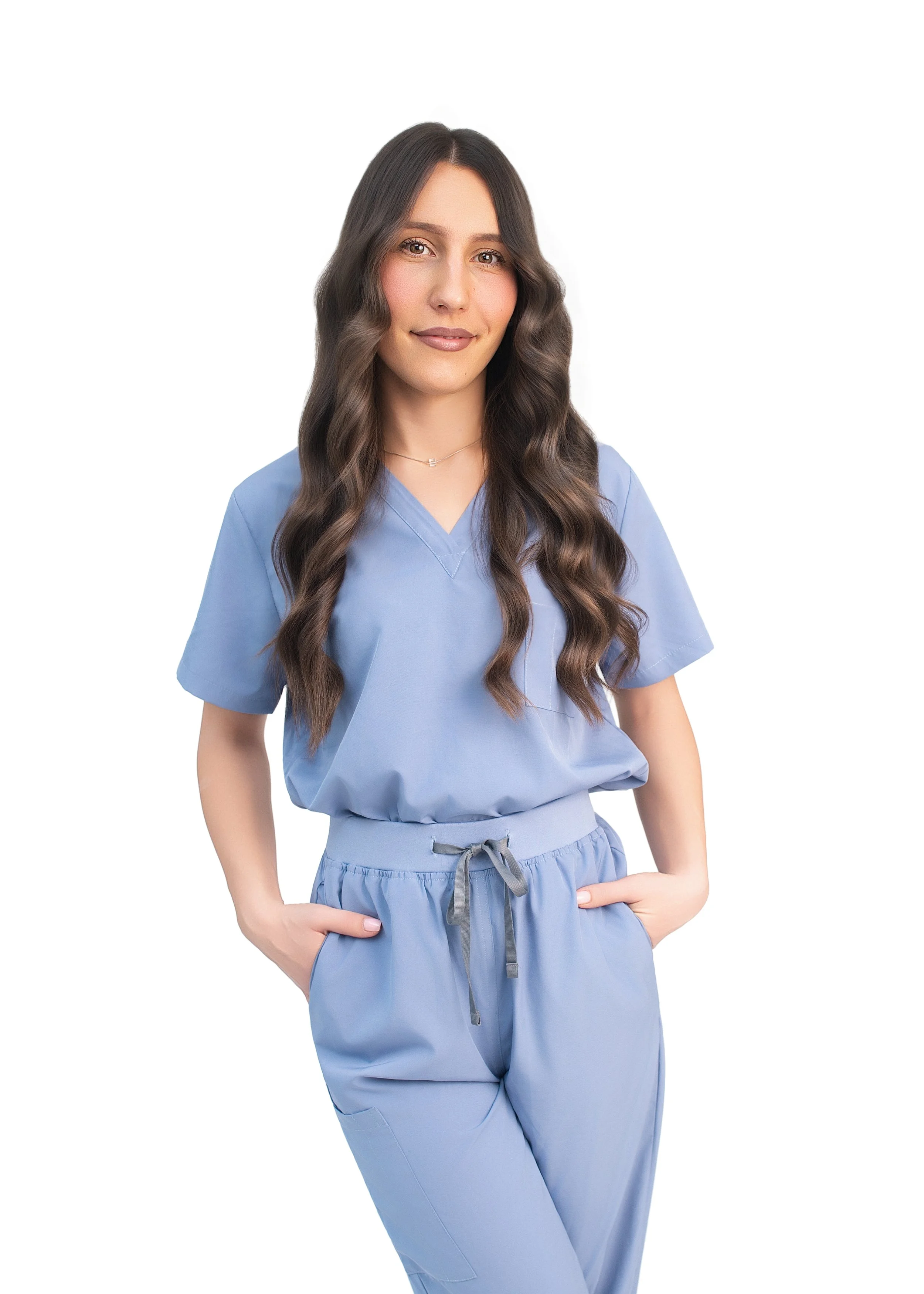 A woman in light blue medical scrubs with long wavy brown hair standing against a white background.