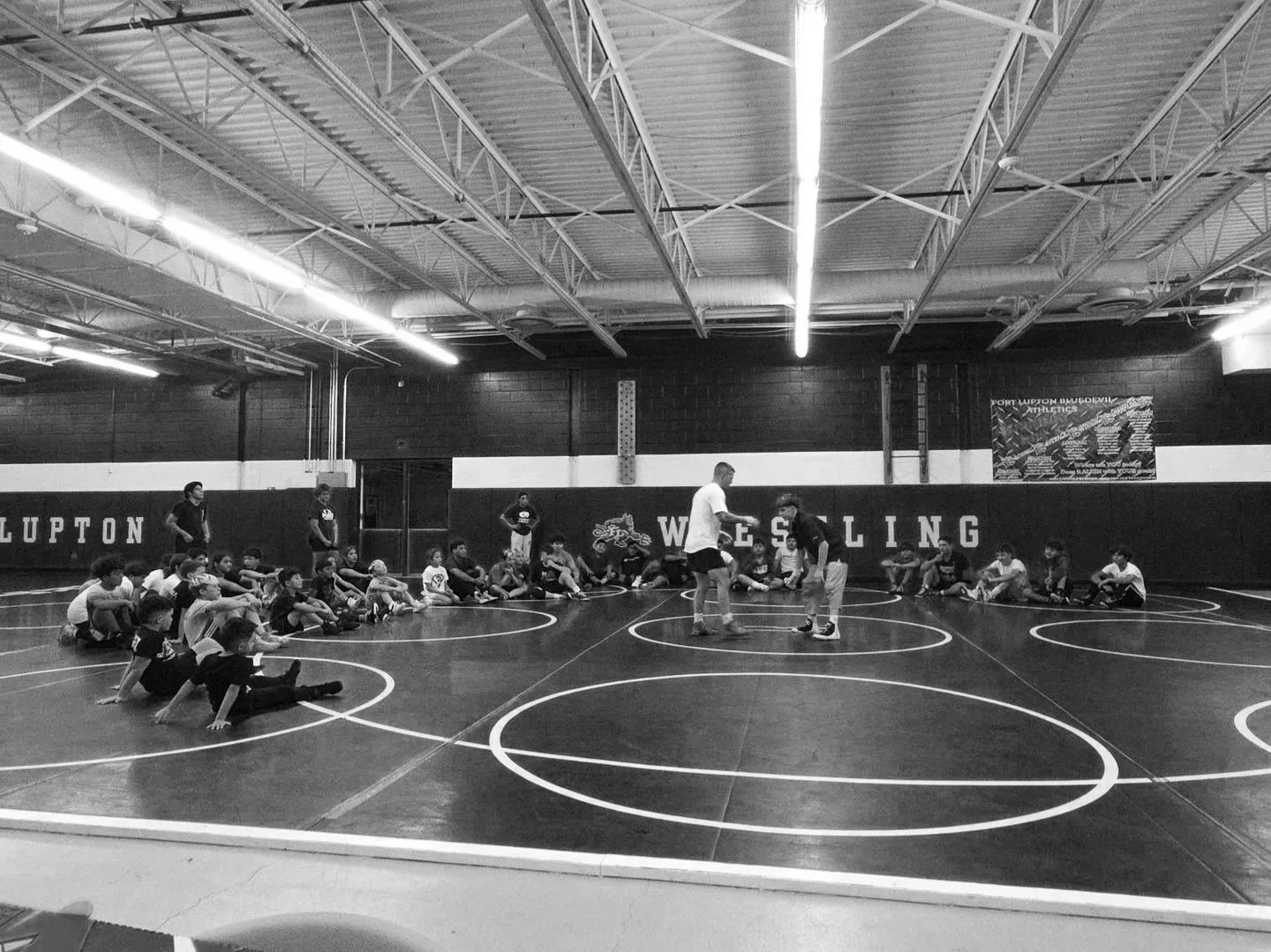 Children sitting on the floor in a gymnasium, watching two boys wrestling, with coaches or instructors standing nearby. The gym has a high ceiling with visible metal beams and a wall with the words 'WRESTLING' painted on it.