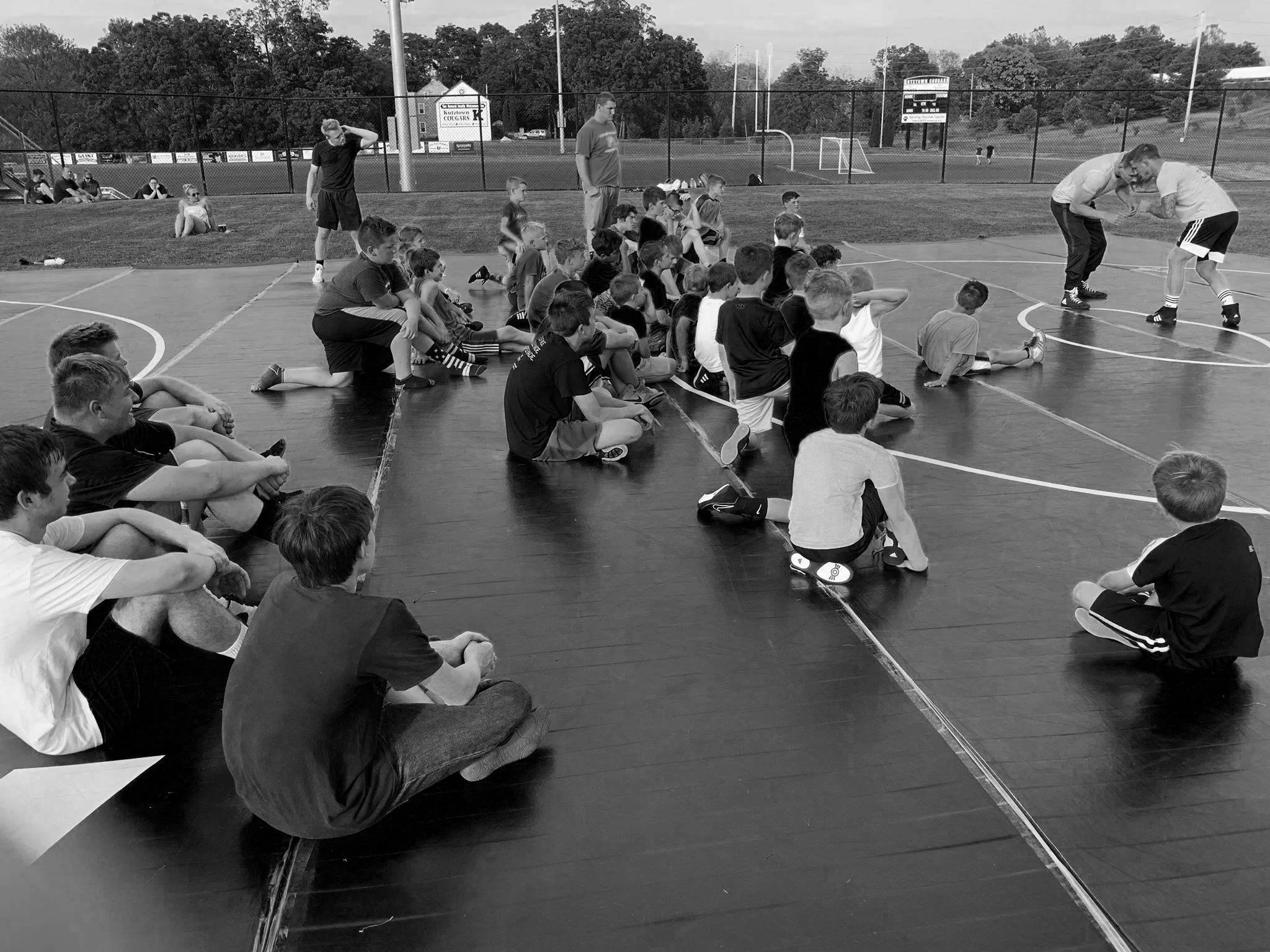 Children seated on the floor of an outdoor basketball court watching two people wrestle.