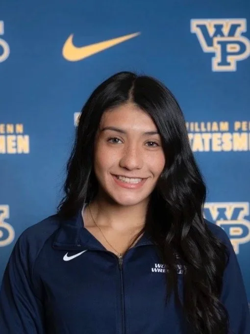Young woman with long black hair smiling in front of a blue backdrop with logos and words, wearing a navy blue sports jacket with a Nike logo.