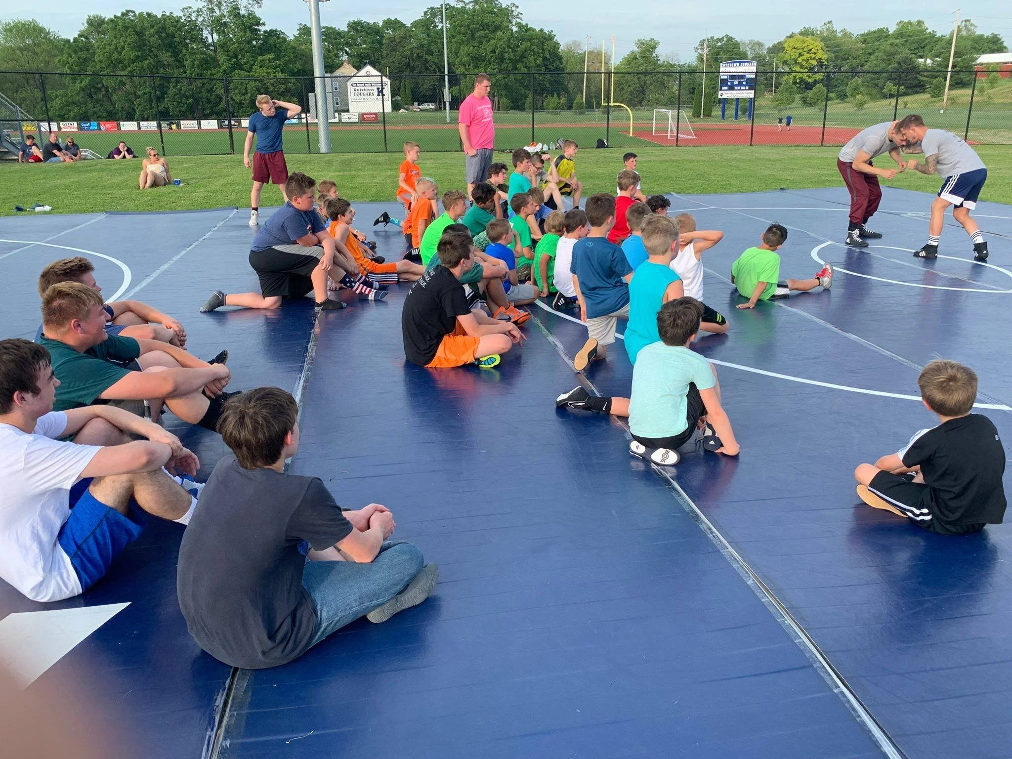 Group of children and teenagers sitting on a blue wrestling mat outdoors at a sports field, watching two wrestlers sparring.