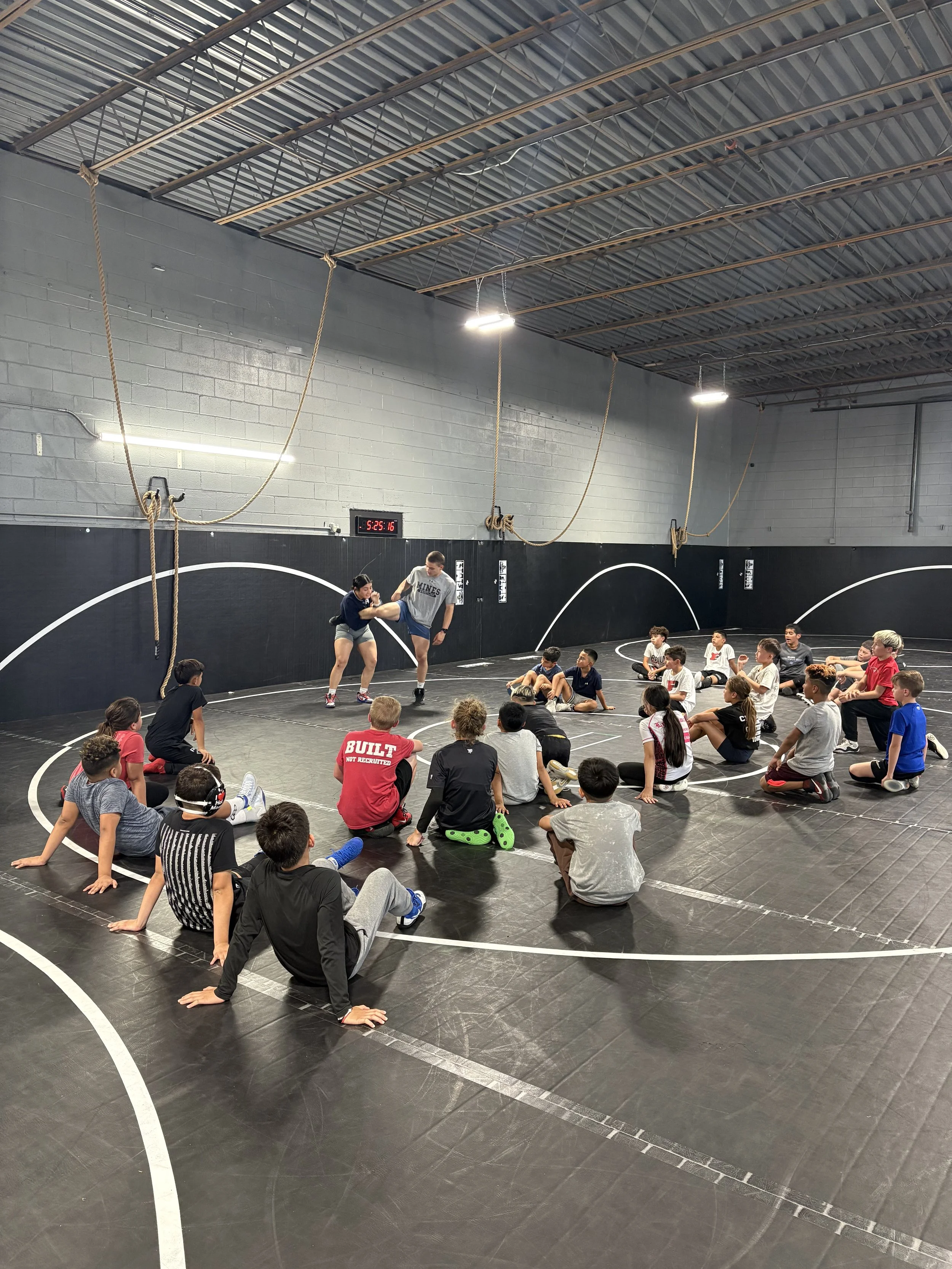 Group of children and teenagers sitting on a blue wrestling mat outdoors at a sports field, watching two wrestlers sparring.