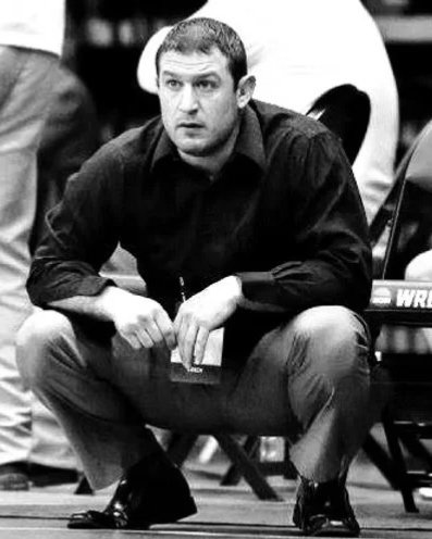 A man with short dark hair, wearing a dark shirt and light-colored pants, sitting on a chair with his arms resting on his knees, in a casual setting.