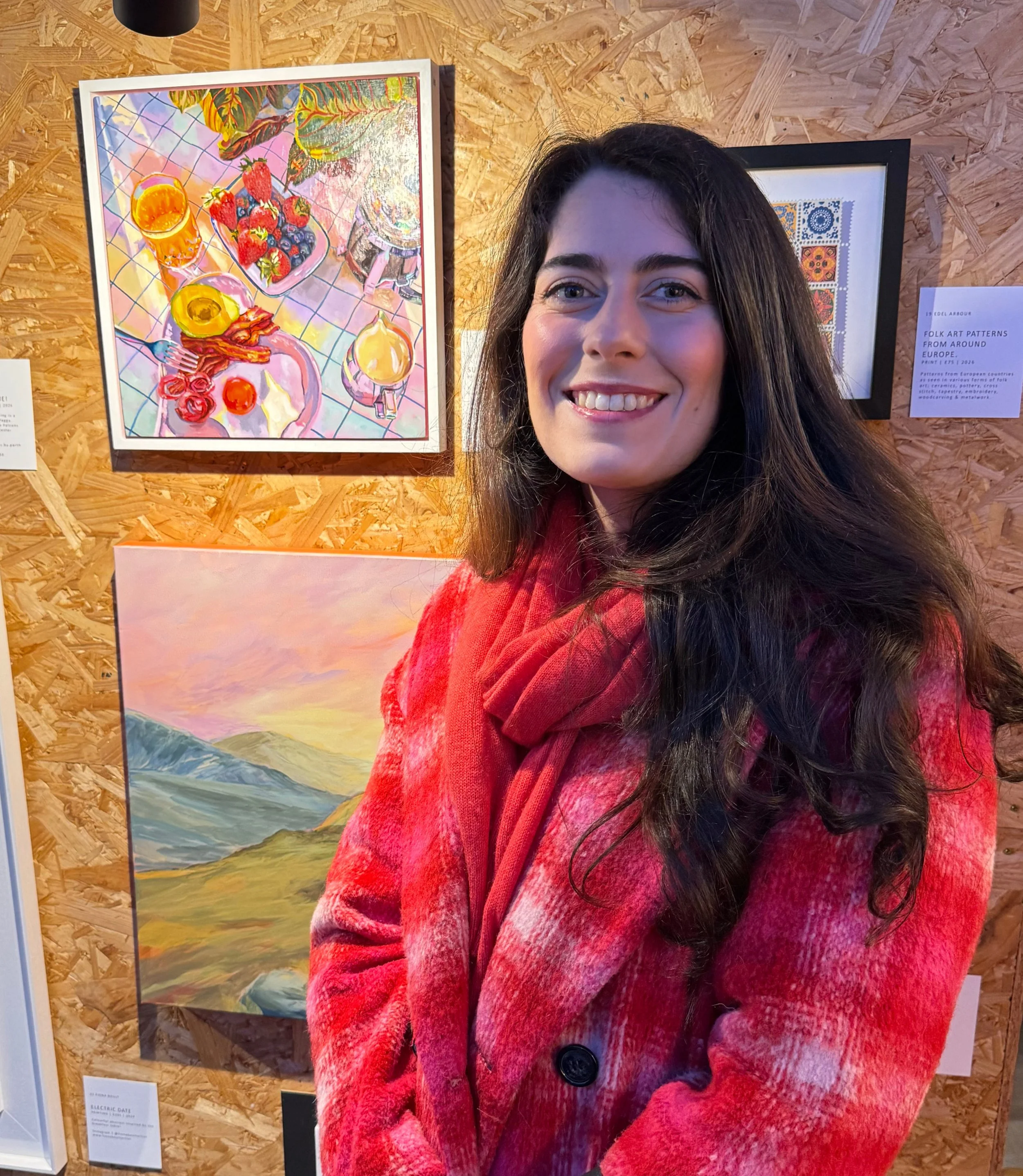The artist "Gabby Nind" a woman with long dark hair smiling in front of colourful artwork displayed on a wooden wall at an art exhibit